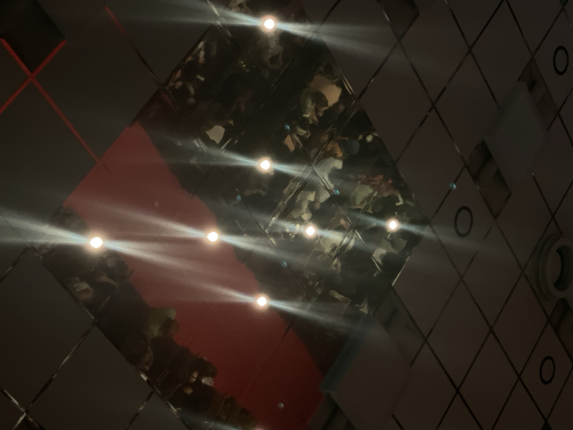 The image is a ceiling view with a mirrored panel in the center reflecting a red wall and people. The mirrored panel is surrounded by dark, square ceiling tiles, some of which have small lights embedded. The overall lighting is dim, with the reflected lights creating streaks of glare. The scene appears to be indoors, possibly in a commercial or entertainment venue. The specific location in Nidda, Germany is not evident from the image itself.