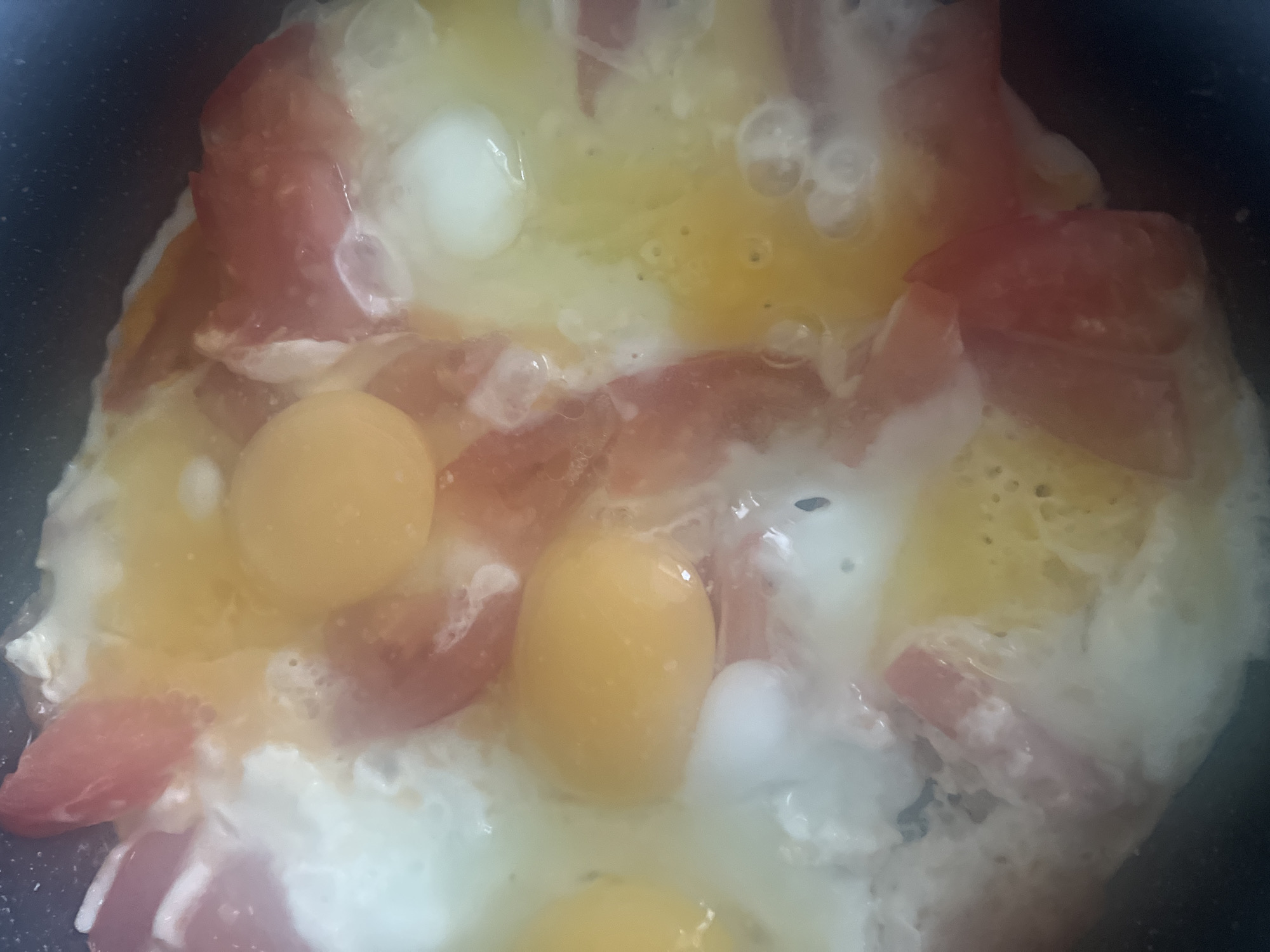 The image shows a close-up, overhead view of a pan containing frying eggs and diced tomatoes. The yolks are bright yellow, and the whites are mostly cooked. The diced tomatoes are red. The scene appears to be indoors. There is no information in the image that would indicate the location is Chaikovsky, Russia, or any information about people, specific actions, or interactions.