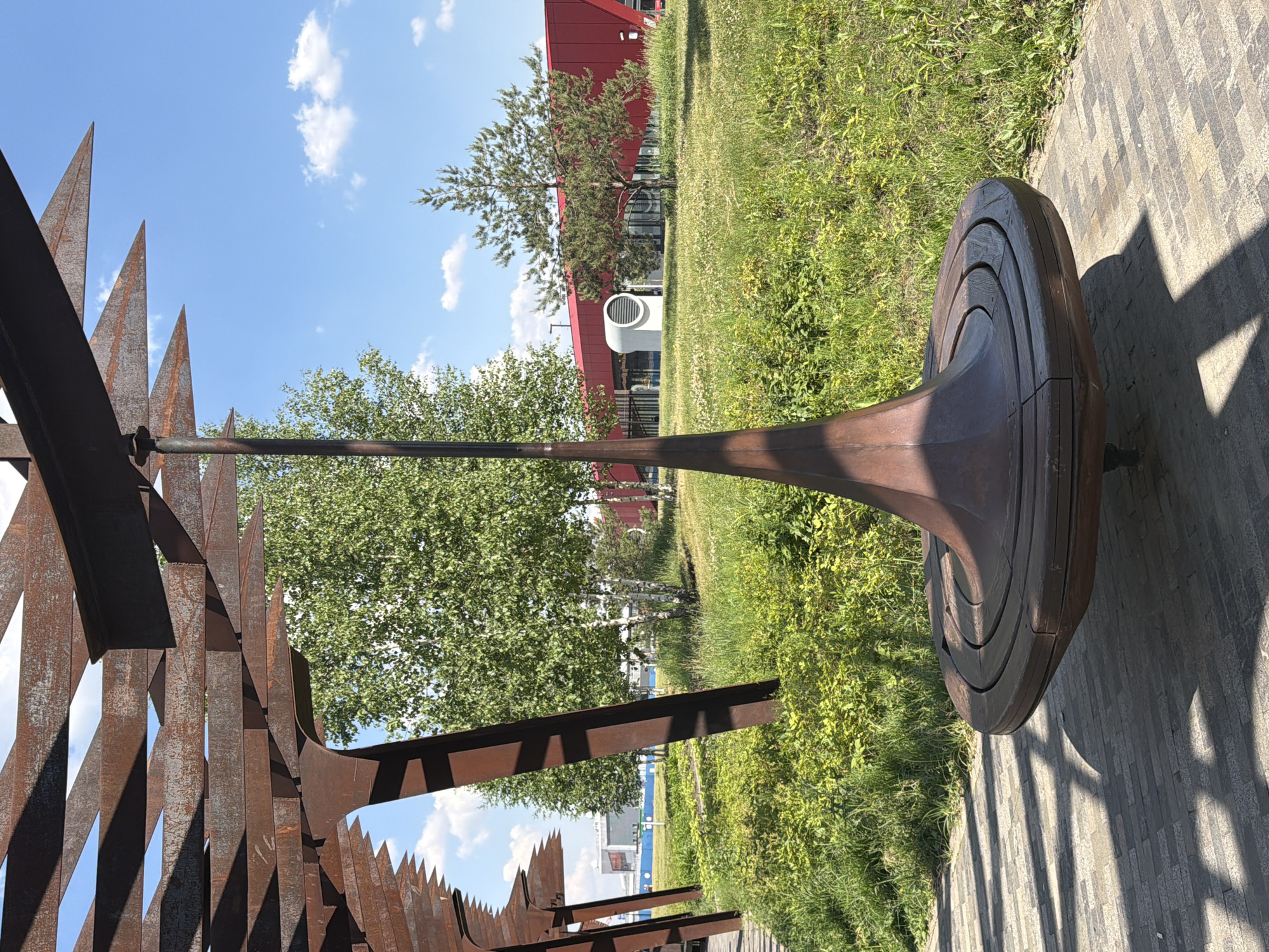 The image depicts an outdoor scene in a park-like setting, likely in Moscow, Russia, characterized by a contemporary architectural design featuring rusty metal structures. In the foreground, a unique, spinning bench made of dark wood and metal is prominently displayed, casting long shadows on the paved walkway. The bench has a conical, central support structure that tapers downwards. Above and to the left, a rusted metal canopy or pergola extends, its geometric design creating an interplay of light and shadow.
Behind the bench, a vibrant green lawn and various trees are visible, including slender birch trees with white bark and a small pine tree. In the background, a modern building with a striking red facade is situated, along with a circular, white ventilation unit on its exterior. The sky is a clear blue with a few scattered white clouds. There are no people or animals visible in the image. The overall impression is one of a modern public space designed with a blend of industrial and natural elements.