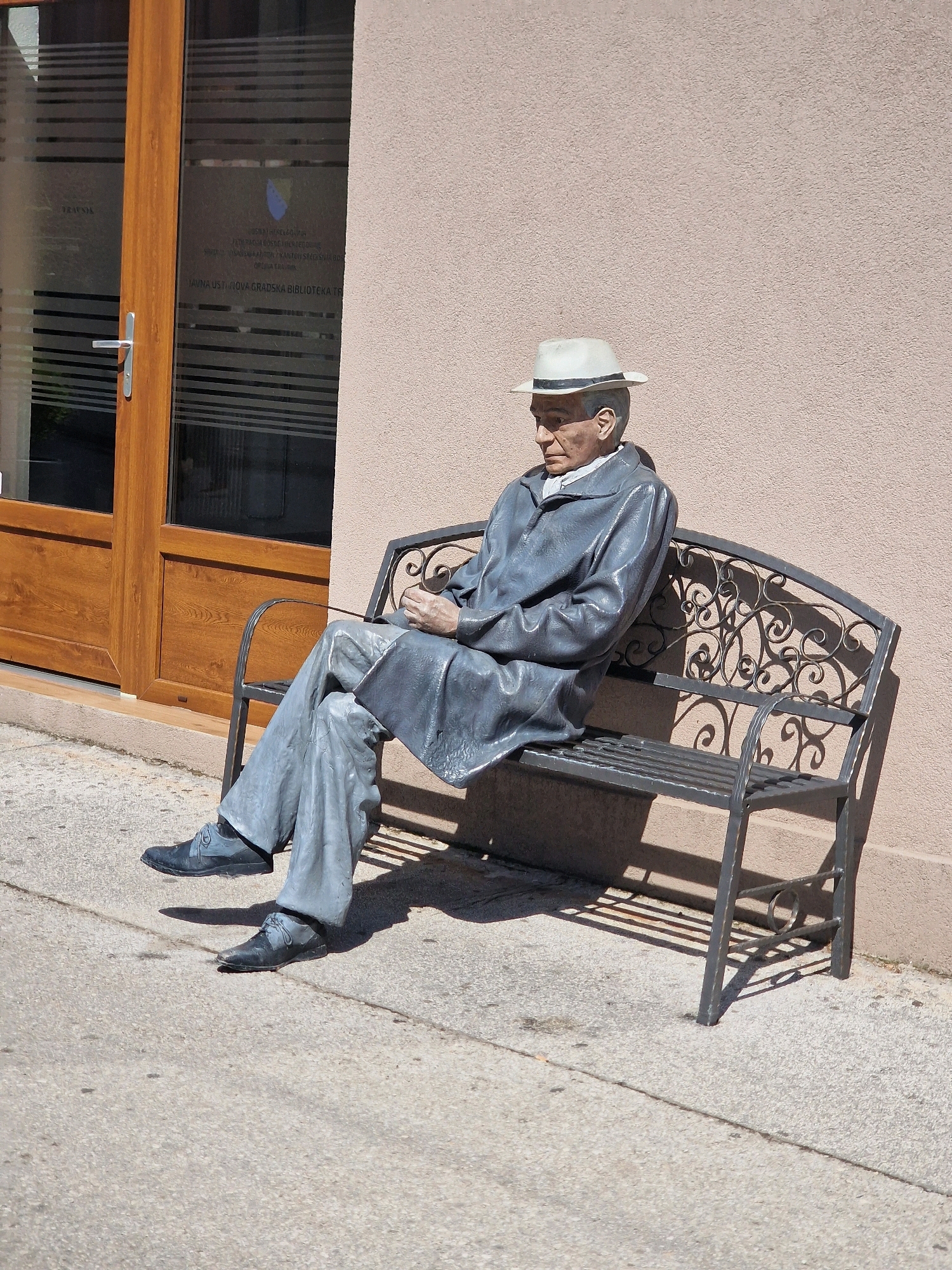The image shows a statue of a man in a hat and overcoat sitting on a decorative metal bench. The statue is positioned on a sidewalk in front of a building with wooden doors and glass windows. Text and a coat of arms are visible on the glass. The location is identified as the Public Institution City Library Travnik, located in Travnik, Bosnia and Herzegovina. The scene is lit by sunlight, casting shadows on the ground and the wall.