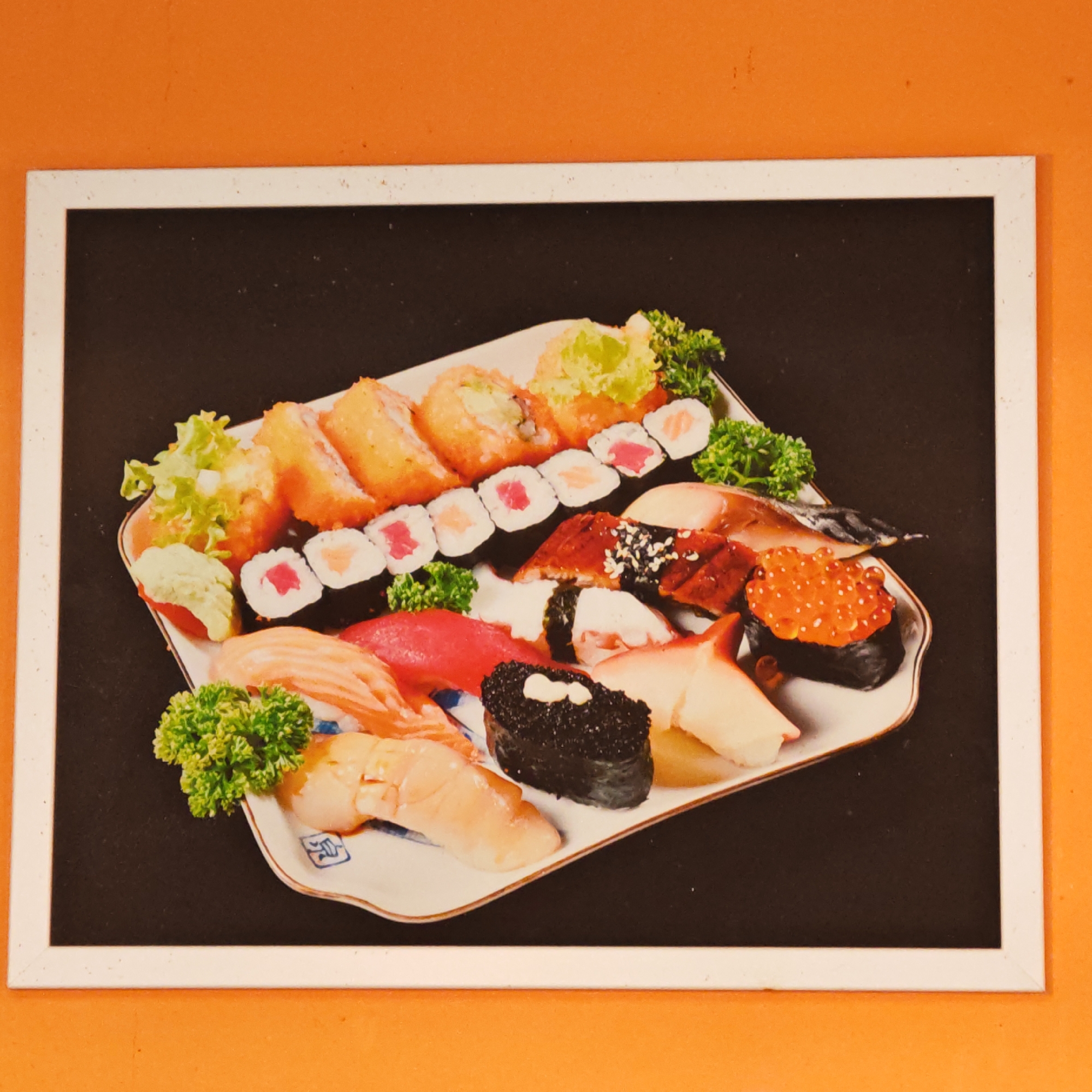 A framed photograph of a plate of sushi is affixed to an orange wall. The frame is white. The photograph depicts a variety of sushi rolls and nigiri arranged on a white plate with blue accents. The background of the photograph is black. The sushi includes various types of fish, vegetables, and roe. There is no visible evidence indicating the city of Rostov, Russia.