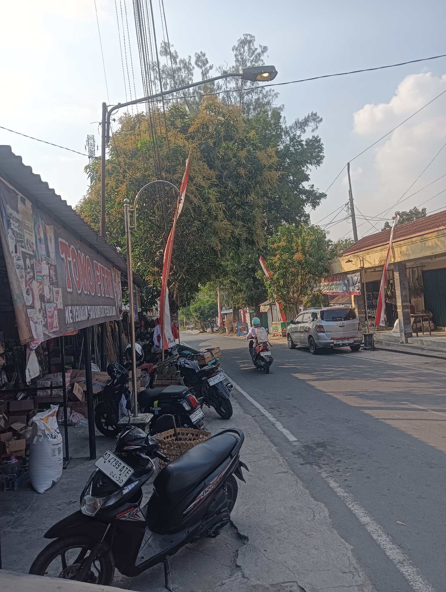 The image depicts a street scene in Palemrejo, Indonesia, under a clear sky with scattered clouds. On the left side of the road, a storefront with a corrugated metal roof displays a banner that reads "TOKO FIRNA." Various items, including bags of merchandise and boxes, are piled near the storefront. Several motorcycles are parked along the sidewalk. In the foreground, a black Honda scooter with license plate "AD 4799 ATE" is prominently featured.
The street itself is paved with asphalt and has a white lane marker. A silver car is driving away from the viewer in the right lane. A person on a motorcycle is also traveling in the same direction. Along the right side of the street are more buildings, one of which has a light-colored facade and a sign above its entrance. Indonesian flags, red and white, are visible on poles in front of some of the establishments. Trees with blooming yellow flowers line the street, and power lines crisscross overhead, supported by utility poles. The overall impression is a typical street view in a residential or commercial area of Indonesia.