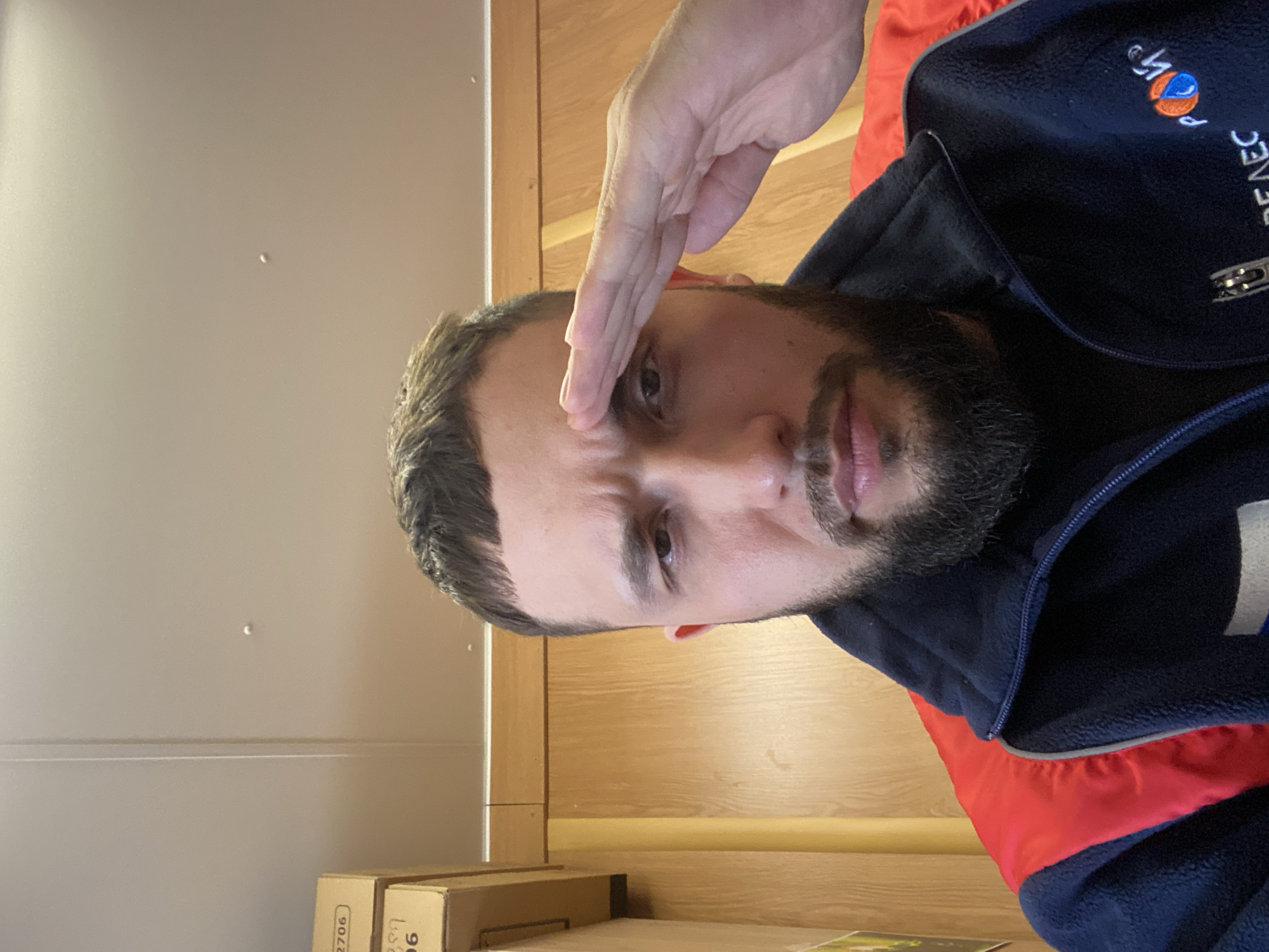 A man with a trimmed beard and short dark hair is shown saluting with his right hand near his forehead. He is wearing a blue fleece jacket with a red accent and a logo on the upper right. The background includes a light-colored wall and wooden panels. Brown cardboard boxes are at the bottom of the frame. The image appears to be taken indoors. The location of Luzhitsy, Russia, is not visibly evident in the image.