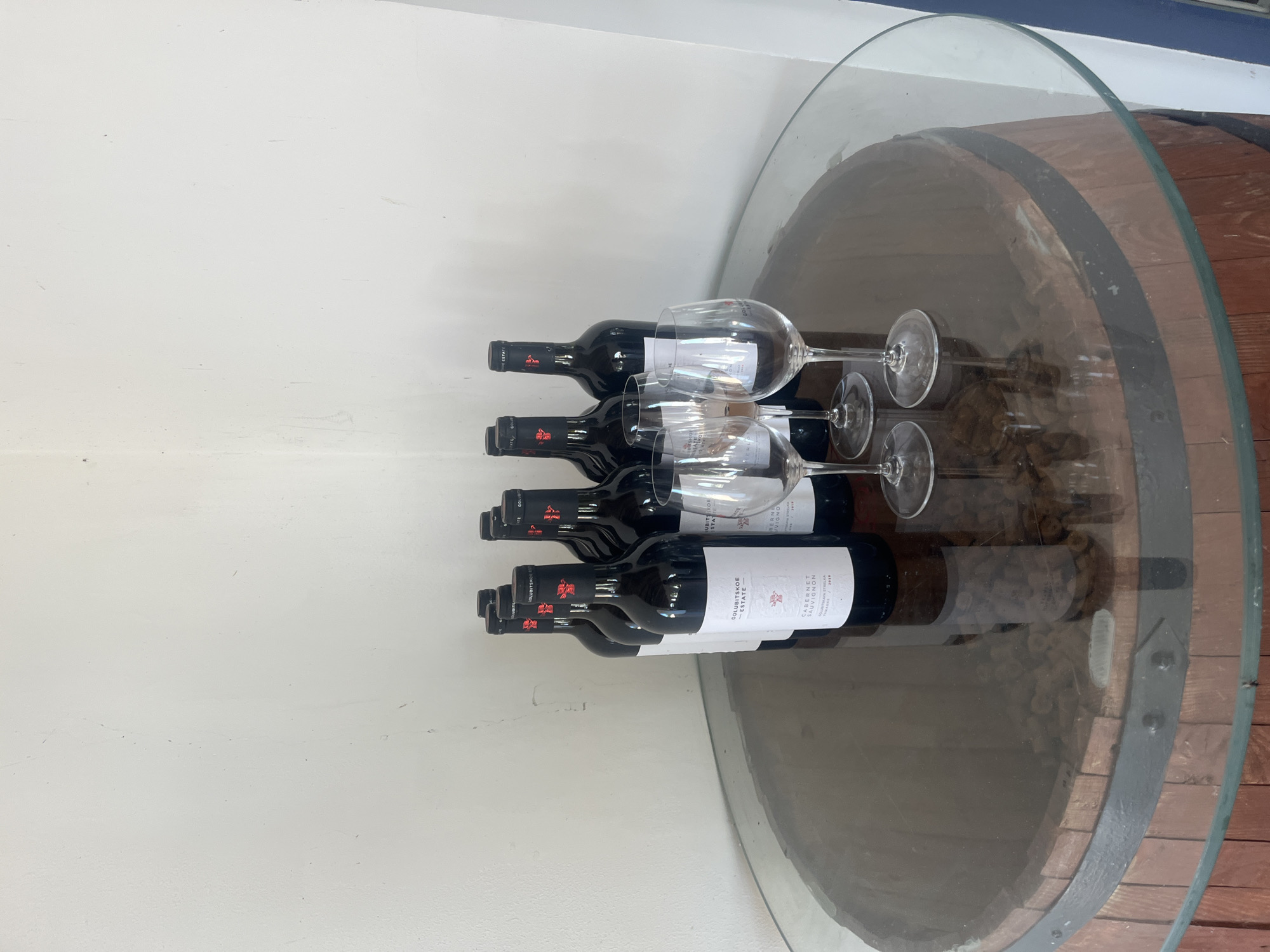 The image displays a tabletop arrangement on a wooden barrel-shaped base, topped with a circular glass surface. On the glass surface, there are eight dark glass bottles of wine, arranged in two rows. In front of the bottles are four clear wine glasses, two of which are filled with a red liquid, presumably wine, and two are empty. The bottles appear to be from "Golubitskoe Estate" and some labels indicate "Cabernet Sauvignon" and the year "2020". The background is a plain white wall. This scene is likely located in Golubitskaya, Russia, given the wine labels.