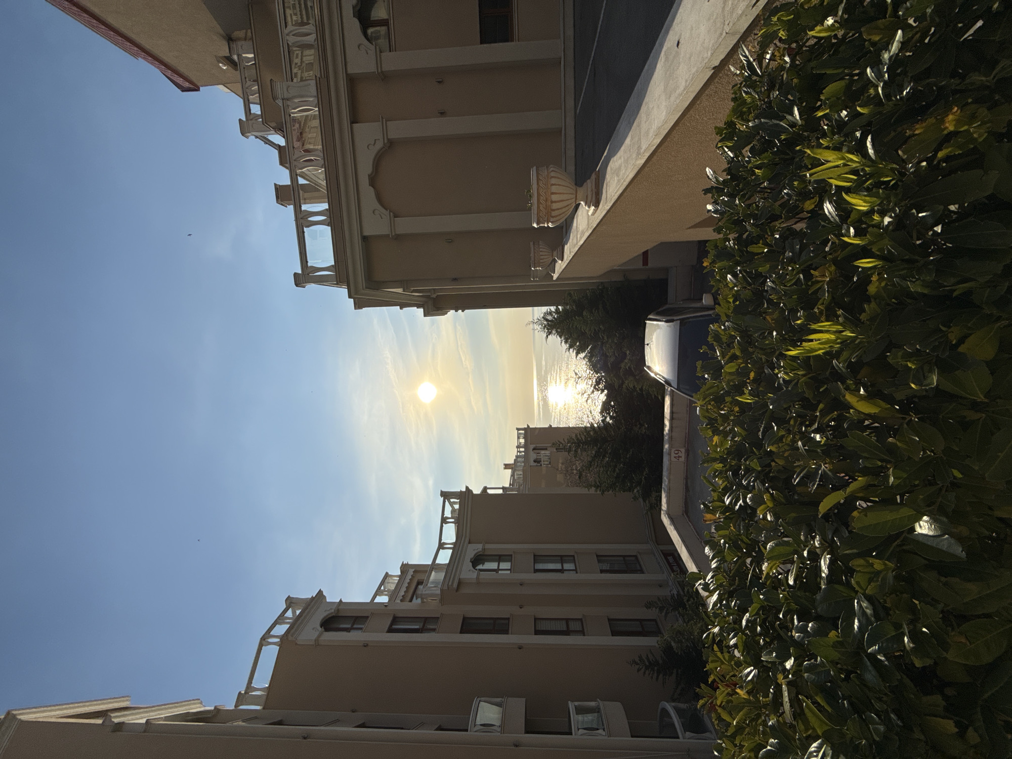 The image depicts a view in Varna, Bulgaria, likely near the coast, based on the sea visible in the distance with the sun reflecting on the water. The perspective is looking down a narrow street or alleyway framed by buildings. The buildings are light beige in color, with architectural details such as balconies with white railings and decorative elements. A bush is visible on the right, partially obscuring a car and a street sign with the number "49". The sky is blue with some wispy clouds, and the sun is visible near the center of the frame, causing a bright glare. There are no people visible. The scene appears to be in daylight, during the late afternoon, with the sun low in the sky.