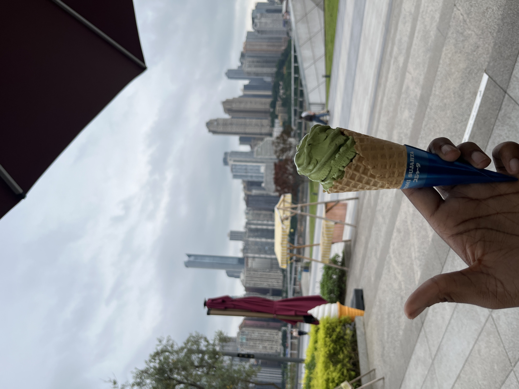 The image depicts a green tea ice cream cone held in a person's hand. The cone is in focus, with the background slightly blurred. The ice cream is a light green color and has a swirled appearance. The cone is a traditional waffle cone. A blue wrapper partially covers the lower portion of the cone.
In the background, a cityscape is visible. The buildings are mostly tall and modern, suggesting a densely populated urban area. The sky is overcast, contributing to a muted color palette.
Other objects in the background include parts of umbrellas, greenery, and structures that appear to be part of the outdoor urban environment. The location appears to be江北城街道, China, evidenced by the buildings and the wrapper on the ice cream cone.