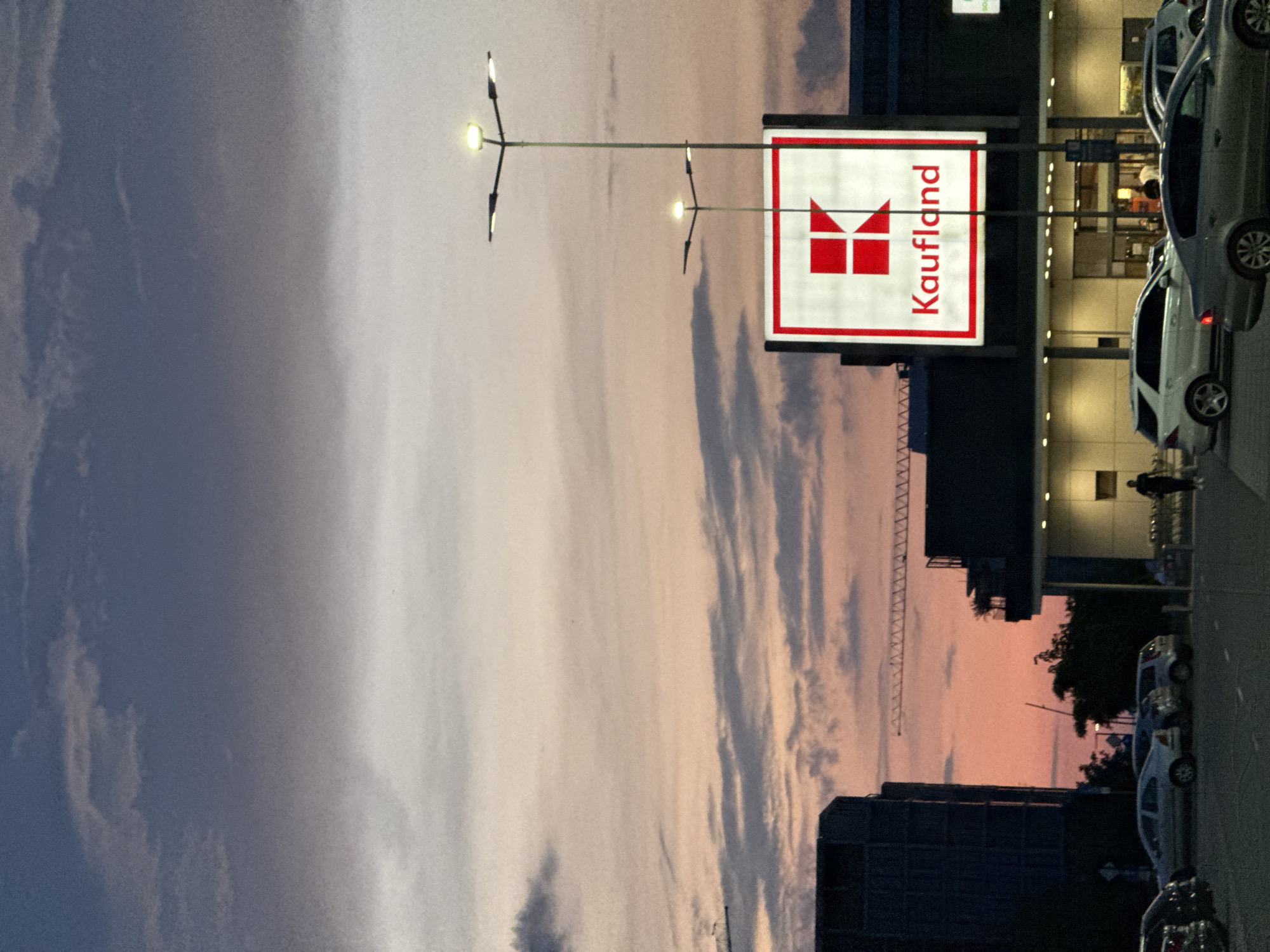 This evening photograph, taken in Burgas, Bulgaria, depicts a commercial area dominated by a "Kaufland" hypermarket at twilight. The sky, visible in the upper and mid-left, transitions from pale grey-blue to soft pink and orange hues, interspersed with long, streaky clouds characteristic of sunset.
Prominently featured in the center-right is a large, illuminated sign bearing the "Kaufland" logo in red text alongside its distinctive red 'K' symbol on a white background. Several modern streetlights with multiple arms and bright, warm-toned bulbs illuminate the immediate vicinity, casting light on the scene.
Below and to the right of the sign, the facade of the Kaufland building is visible, with a well-lit entrance suggesting ongoing activity. A silver sedan and a white SUV are among several cars parked in a lot or on the street in the foreground on the right. At least two individuals are present: one person is seen walking near the store entrance, and another is partially visible further down among the parked vehicles.
In the mid-left distance, a large, dark structure resembling a construction crane extends horizontally across the frame, silhouetted against the colorful sky. Other darker buildings are also visible in the background. The scene conveys a calm, routine evening atmosphere in an urban setting.