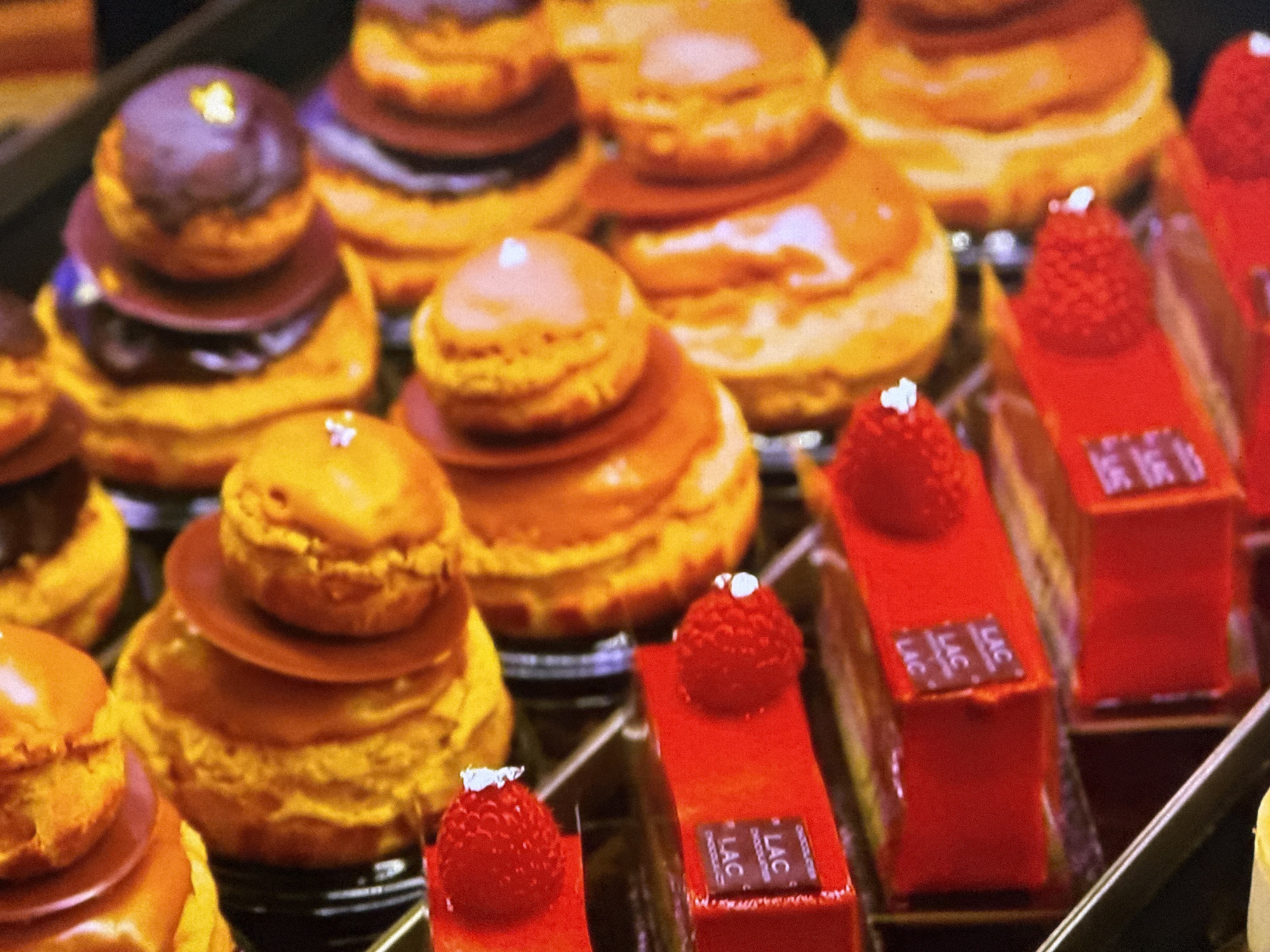 A display case shows two types of pastries. One type consists of cream puffs stacked into small towers with a chocolate glaze or a caramel-like glaze, and a small piece of foil on top. The other type is a rectangular slice with a red glaze and a raspberry on top. Each slice has a small square of chocolate with the logo "LAC" on it. The location is likely a patisserie in Valencia, Spain, based on the pastries' style and appearance.