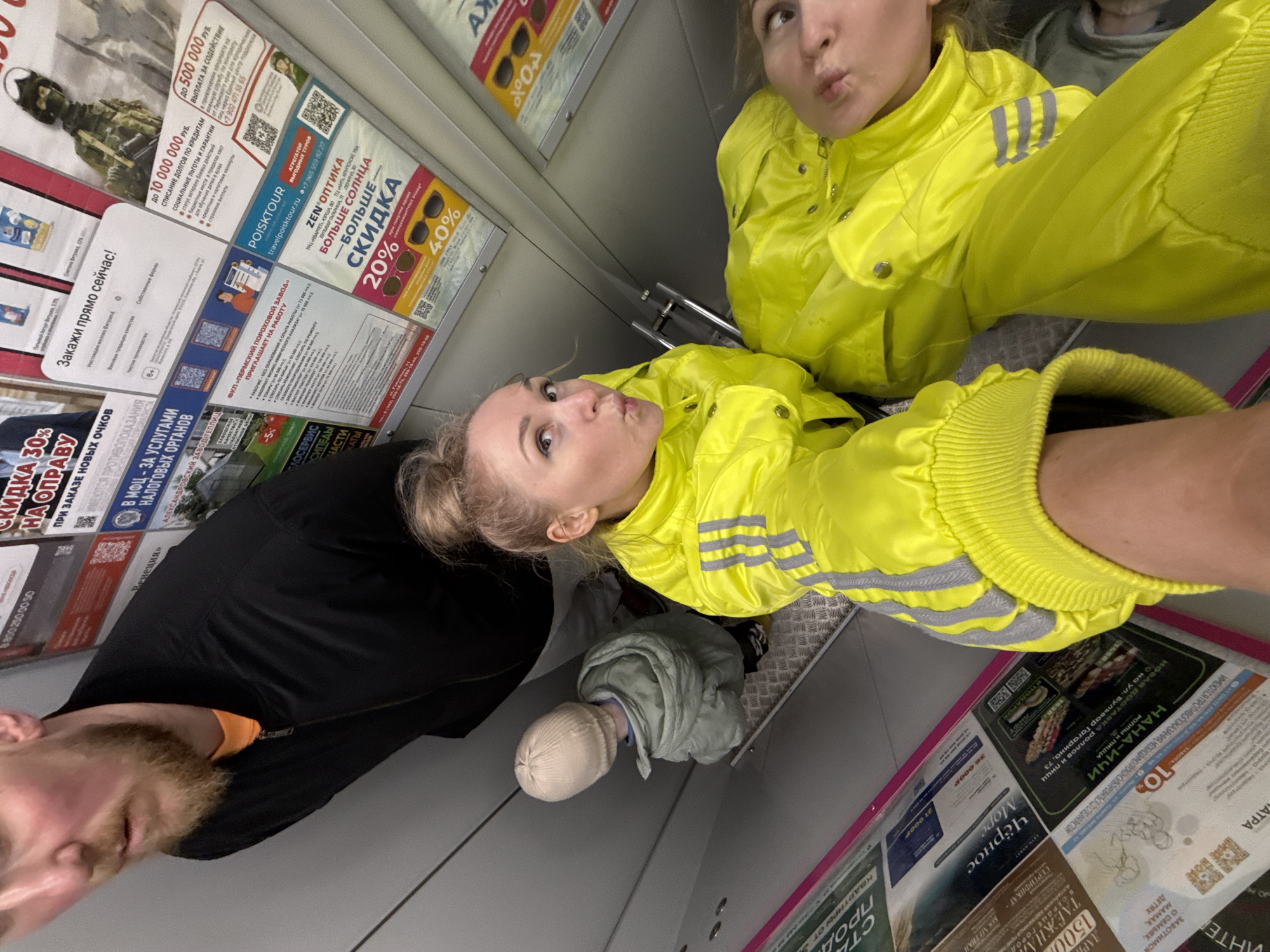The image shows a group of people in an elevator in Perm, Russia. Two women in yellow jackets are making faces at the camera. A man in a black jacket is partially visible in the lower left corner. A child in a light green jacket and beige hat is standing in the middle.
The elevator walls are covered in advertisements, including those for Poistour travel agency, Zen Optika, and a job posting for the Perm Powder Plant. Other ads are visible for local businesses, like Nana-Ichi pizza and sushi, and for apartments. The Zen Optika ad offers a 20% discount and mentions locations on Gagarin Boulevard and Lenin Street in Perm.
