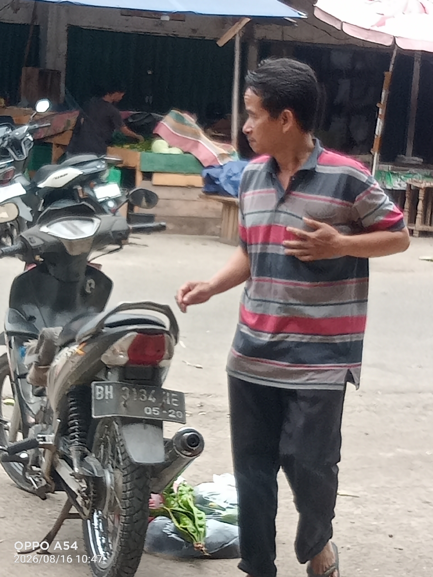 An Indonesian man in a horizontally striped polo shirt and dark pants stands by a parked scooter in what appears to be a market. The scooter's license plate reads "BH 3134 ME 05-20". In the background, a person in a dark shirt is visible behind a stall with various goods, including what looks like produce wrapped in plastic. The environment suggests an outdoor market setting, possibly in the city of Jambi, Indonesia, given the license plate format. The man is looking to his left with his right hand resting on his chest, and his left arm is extended.