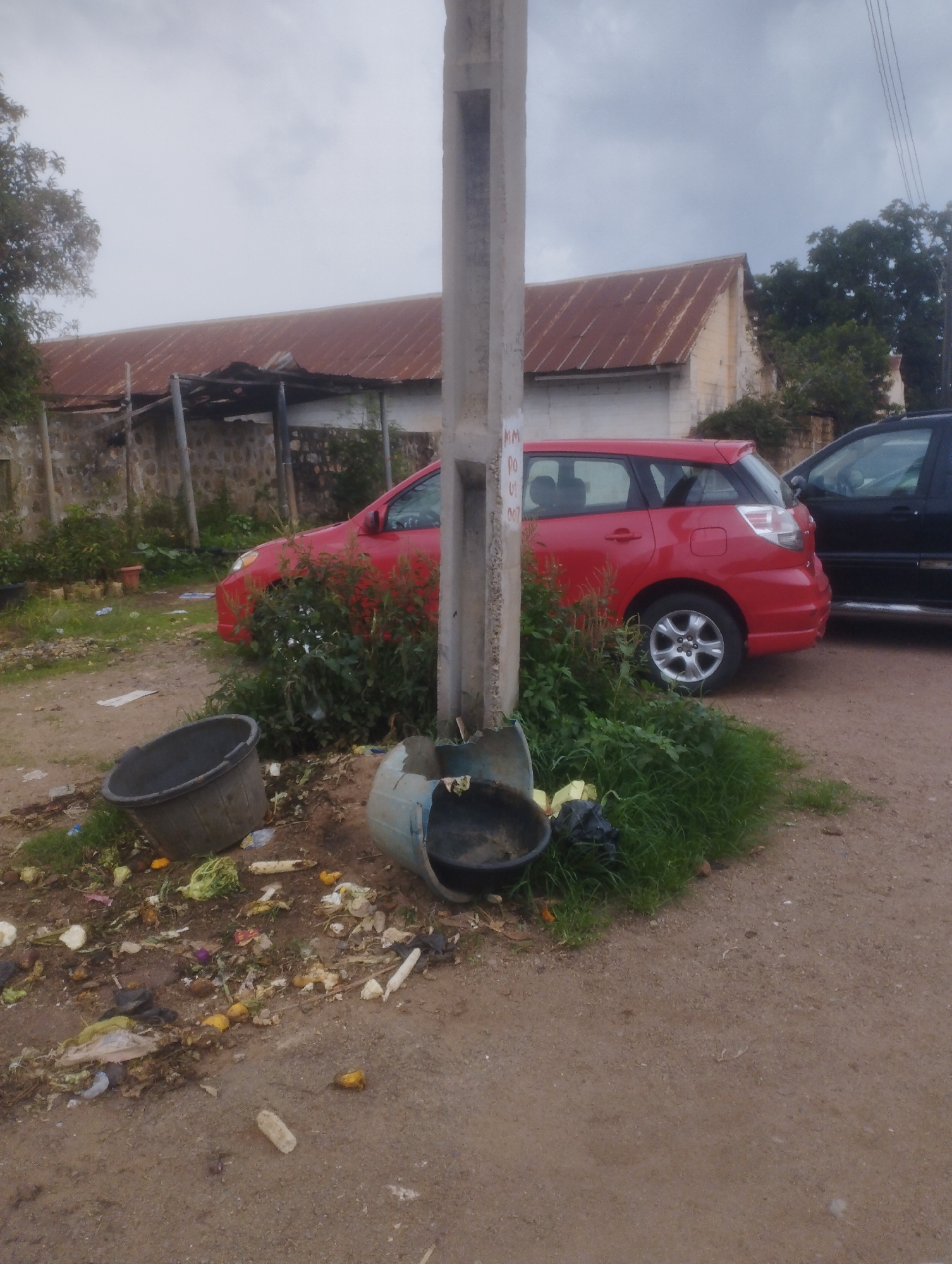 This image captures an outdoor scene, likely a roadside or an unpaved area in Jos, Nigeria, under an overcast sky. The foreground is dominated by unpaved dirt ground, extensively littered with organic waste, including vegetable scraps, fruit peels, and corn cobs, alongside some plastic and paper debris. Two dark-colored plastic buckets are visible on the ground; one is intact but tipped over, and the other is broken into several pieces, contributing to the scattered refuse.
A prominent concrete utility pole stands slightly off-center in the midground, partially obscuring a red hatchback car (possibly a Toyota Matrix) parked behind it. Overgrown green bushes and weeds sprout around the base of the pole and the front of the red car. To the right of the red car, the front portion of a dark-colored SUV or truck is also visible.
In the background, a building with a rusty, corrugated iron roof and light-colored walls can be seen. A stone wall or fence runs along the perimeter of the property behind the parked cars and bushes. Trees are visible beyond the building on the right side of the frame.
There is no direct human activity depicted. The visible elements suggest a general street or informal area where waste may be accumulating. On the concrete utility pole, faint handwritten black text is visible, including "MM" and partial numbers or letters like "DO", "01", and "007", which appear to be unofficial markings. The daytime setting is indicated by the light, though the heavy cloud cover gives a muted illumination.