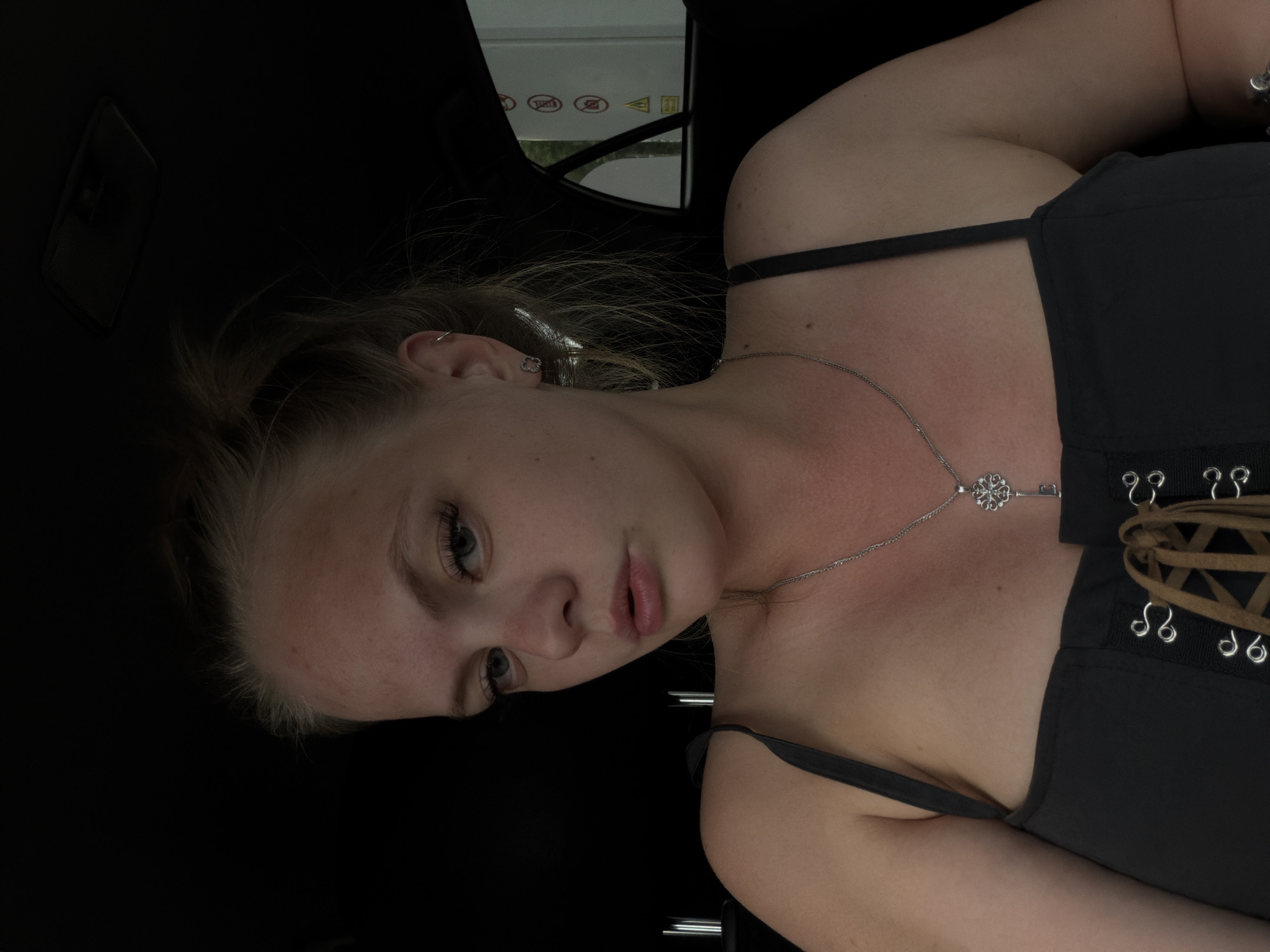 A young woman with blonde hair tied up is sitting in a car. She is wearing a black tank top with lacing at the front and a silver necklace with a key pendant. She has on small stud earrings and a hoop earring in her other ear. The interior of the car is dark, with the exception of some light coming in from the side window, revealing a glimpse of a building with red and white warning signs. The photo appears to have been taken in Chaikovsky, Russia, as suggested by the presence of a building with warning signs that are common in industrial or public areas.