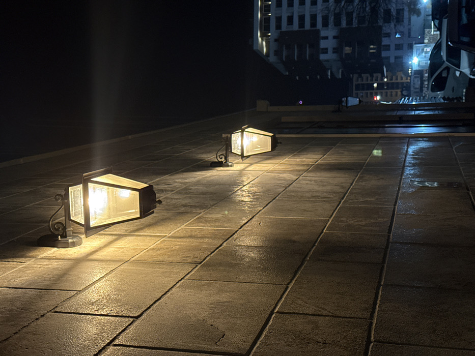 The image depicts an urban nighttime scene in Budva, Montenegro. The primary focus is a light brown-colored building wall, appearing to be constructed of large rectangular blocks or bricks with visible mortar lines. Two illuminated wall-mounted lanterns are affixed to this wall. The upper lantern, positioned towards the upper-left, is closer to the viewer, brighter, and casts a prominent upward-diagonal beam of warm white light across the textured wall surface. This lantern has a dark-colored, traditional-style frame, likely hexagonal or octagonal, with clear glass panels. A second, identical but smaller lantern is mounted further down and to the right on the same wall, also illuminated.
To the right of the wall, a street or parking area is visible, occupied by at least three parked vehicles, including a white sedan, a dark-colored vehicle, and a light blue/grey vehicle. Distant artificial illumination from streetlights and windows of multi-story modern buildings is visible against a dark night sky. One prominent background structure is a tall building with multiple lighted windows. A leafless tree is faintly discernible between the street and the distant architecture. No individuals are present in the observable scene.