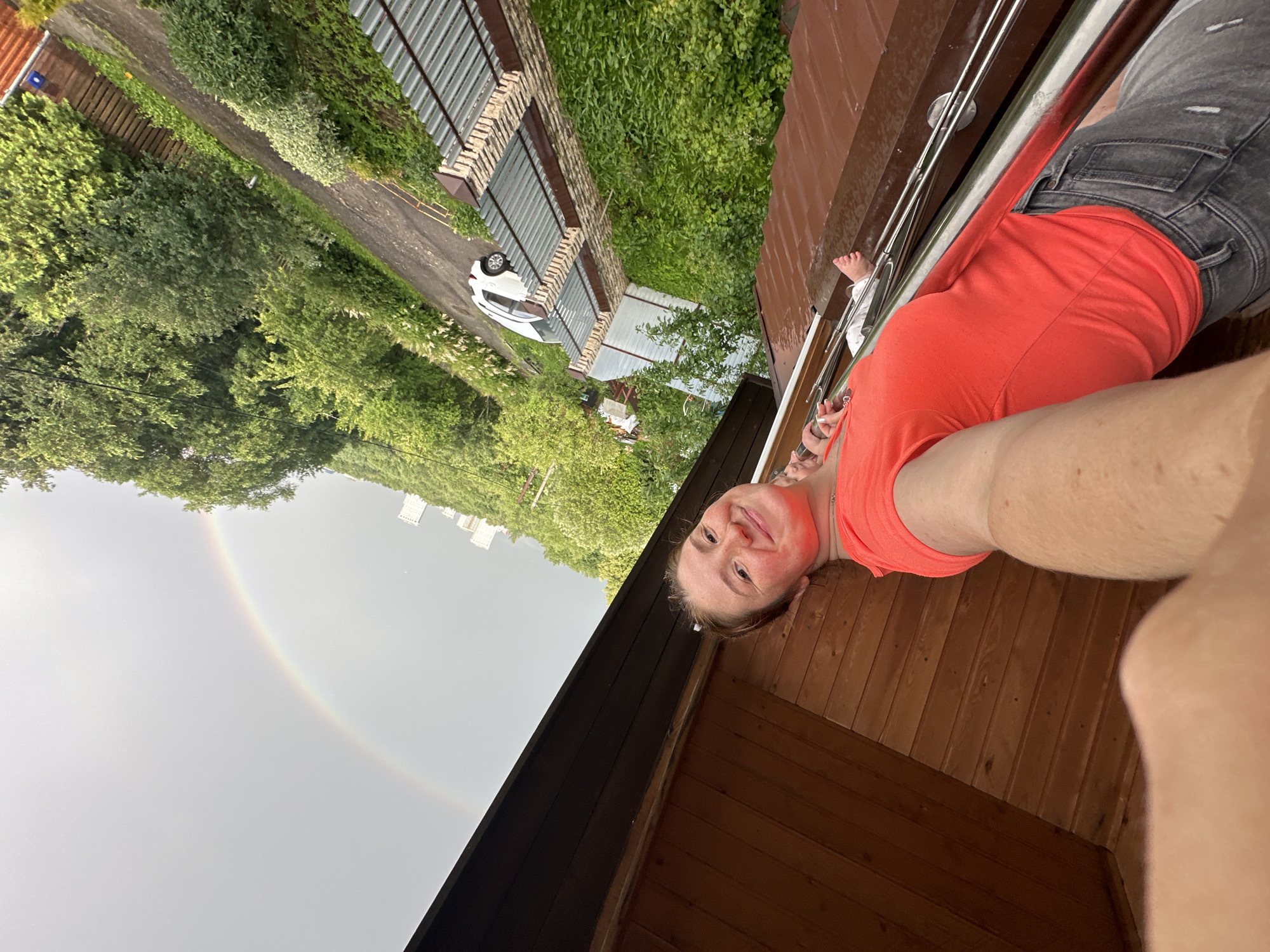 In Nizhny Novgorod, Russia, a young woman with fair skin and freckles smiles at the camera, taking a selfie from a low angle. She is wearing a bright orange t-shirt and grey jeans. Her left arm is extended, holding the camera. Her right arm is bent at the elbow, resting on a wooden surface that appears to be part of a balcony or roof. In the background, a faint rainbow arcs across a cloudy, grey sky. Beyond the rainbow, several tall, white buildings are visible, suggesting an urban setting. Lush green trees and foliage line a pathway or road to the right of the frame, and a white car is parked along it. The scene appears to be outdoors, possibly after a rainfall, given the subdued lighting and the presence of the rainbow.