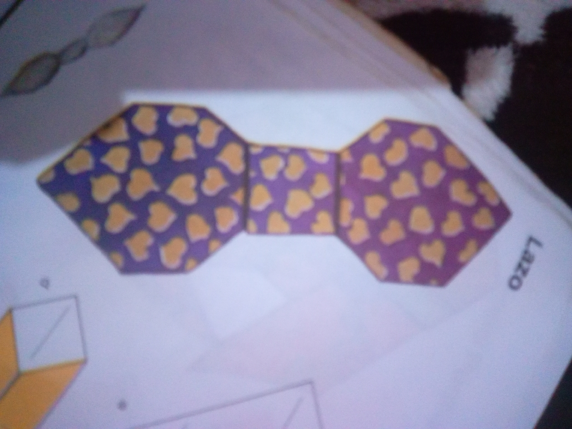 The image shows a close-up, slightly blurry view of a purple bow tie pattern with yellow heart shapes. The bow tie is made of folded paper. The bow tie is placed on a white surface, possibly a piece of paper. To the right of the bow tie, the word "LAZO" is visible, written vertically and upside down. In the lower left corner, a partial view of a geometric drawing, possibly a cube or prism, is visible. The background appears to be a desk or table, with a dark, fluffy object (perhaps fabric or fur) in the upper right corner. There is no indication of the city of La Tagua, Venezuela, or any people in the image. The scene is a static, still life arrangement.