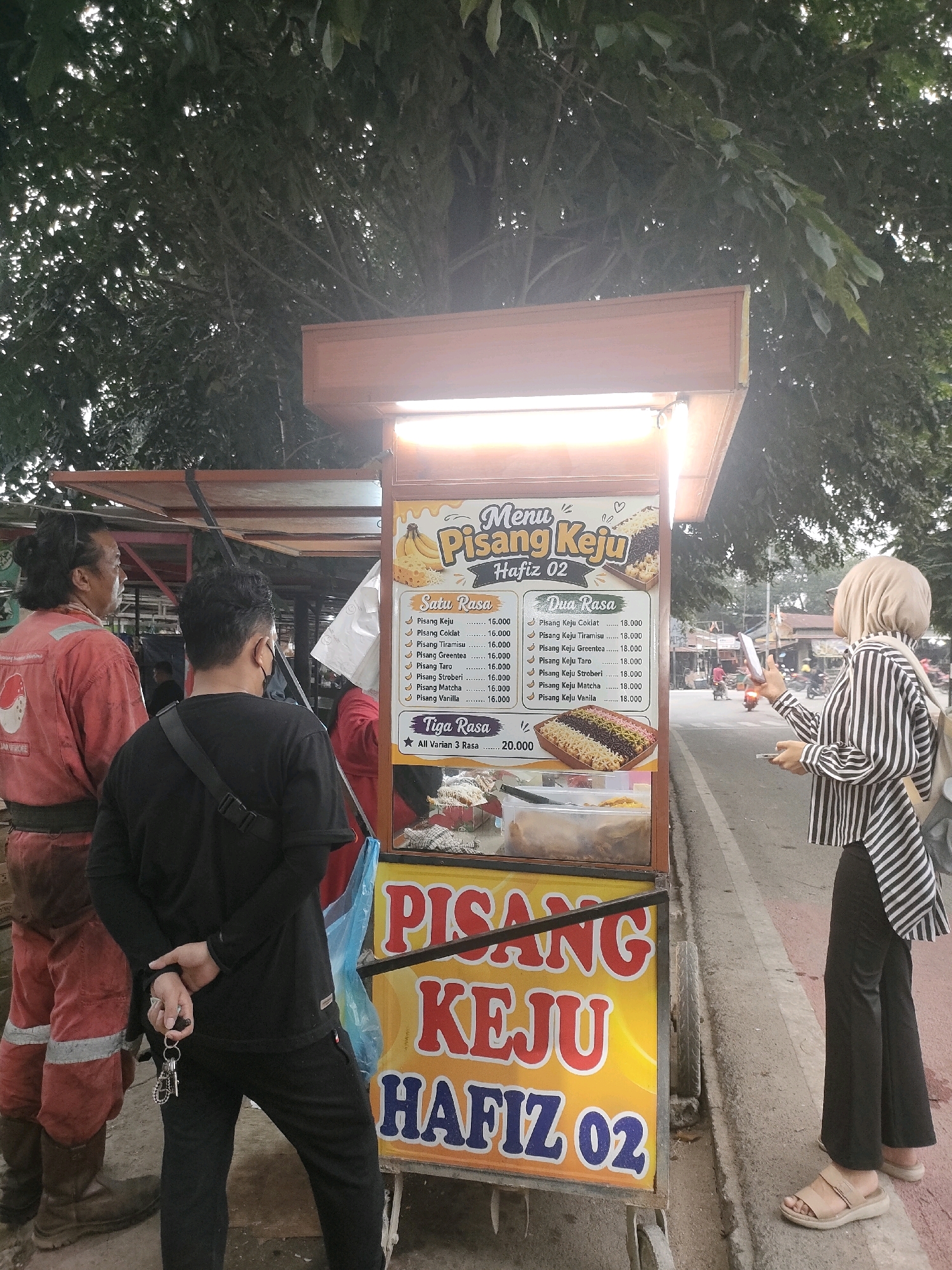 A street food cart selling "Pisang Keju" (cheese banana fritters) is situated on the side of a road in Batam, Indonesia, likely in a commercial or residential area with moderate foot traffic. The cart features a prominent sign with the brand "Pisang Keju Hafiz 02" and a menu listing various flavors. A man in a red uniform with white stripes is standing to the left, facing the cart. Next to him, a man in a black t-shirt and black pants, holding keys, is also facing the cart. To the right, a woman wearing a striped long-sleeved shirt and a headscarf is standing, holding a mobile phone and a small bag. The background shows trees and a street with some passing vehicles and buildings. The lighting suggests it is daytime, possibly late afternoon, with some dappled sunlight filtering through the trees.