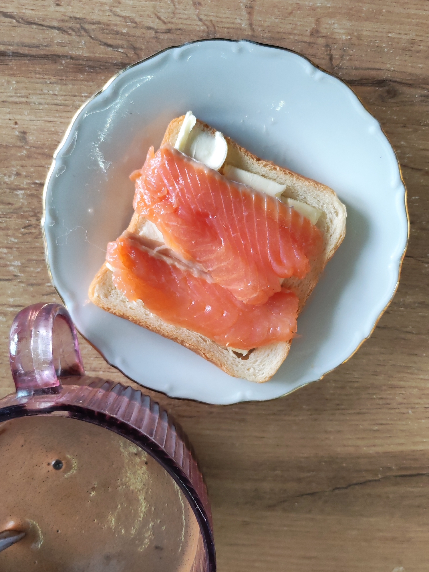 A top-down shot captures a breakfast or light snack set on a wooden table in Izhevsk, Russia. The main focus is a single slice of white toast, generously topped with multiple slices of bright orange, smoked salmon. Several small, pale yellow pats, likely butter or cream cheese, are visible nestled amongst and on top of the salmon. This meal is served on a delicate, white porcelain plate featuring a scalloped edge and a thin gold rim. To the bottom left of the plate, a portion of a pink, translucent glass mug with a ribbed texture is visible. The mug contains a frothy, light brown beverage, which appears to be coffee or a similar drink. The wooden table surface, with its distinct grain pattern, serves as a natural and warm backdrop for the meal. The lighting is bright and consistent, suggesting an indoor setting, possibly a home kitchen or dining area. No text is visible in the image.