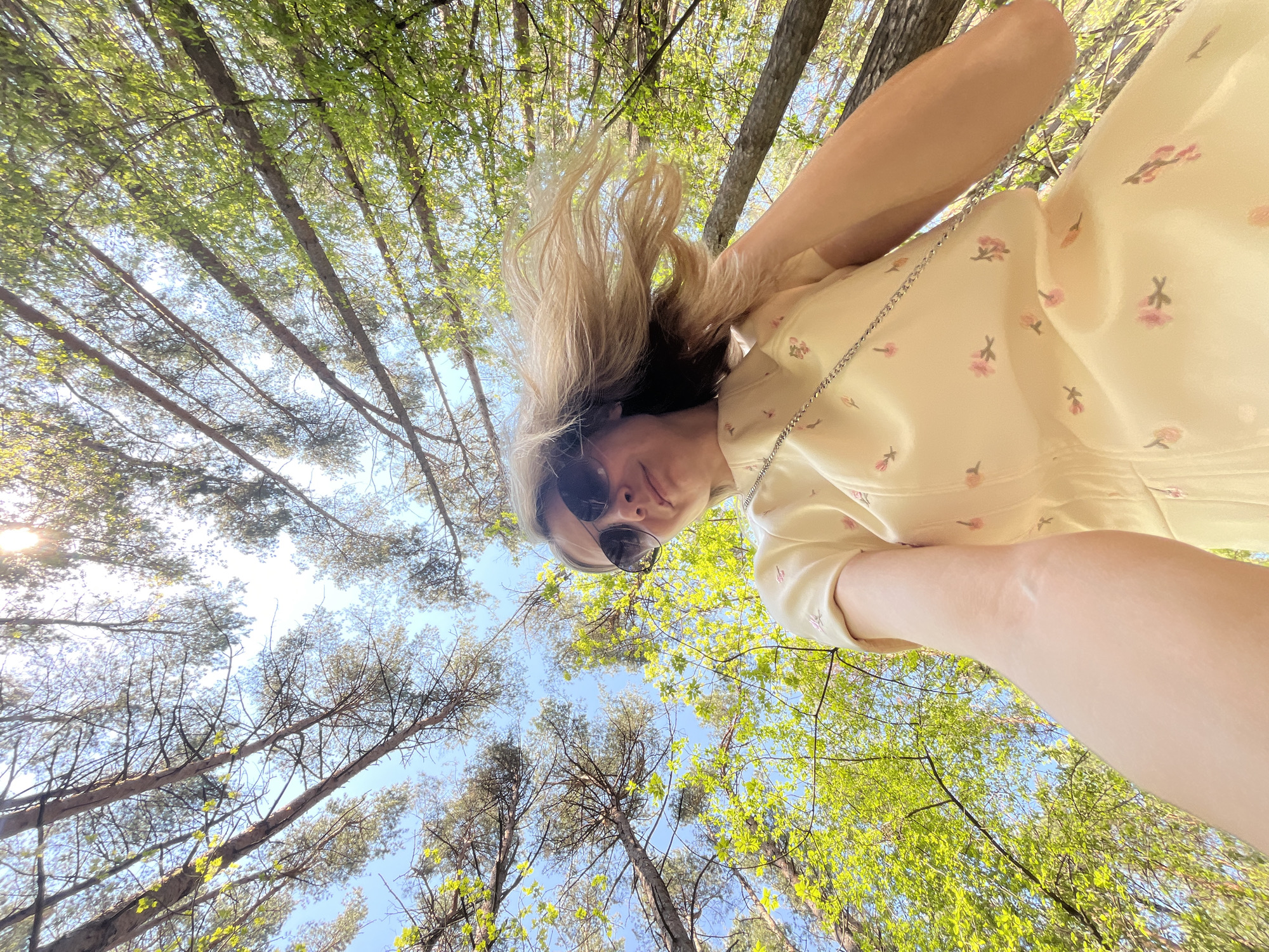 A low-angle shot taken from beneath the subject, showcasing a person wearing sunglasses and a light-yellow floral dress, positioned against a backdrop of tall trees in the city of Chaikovsky, Russia. The sky is visible through the canopy of leaves, suggesting a sunny day. The focus is on the person's face and upper body, with the trees framing the scene.
