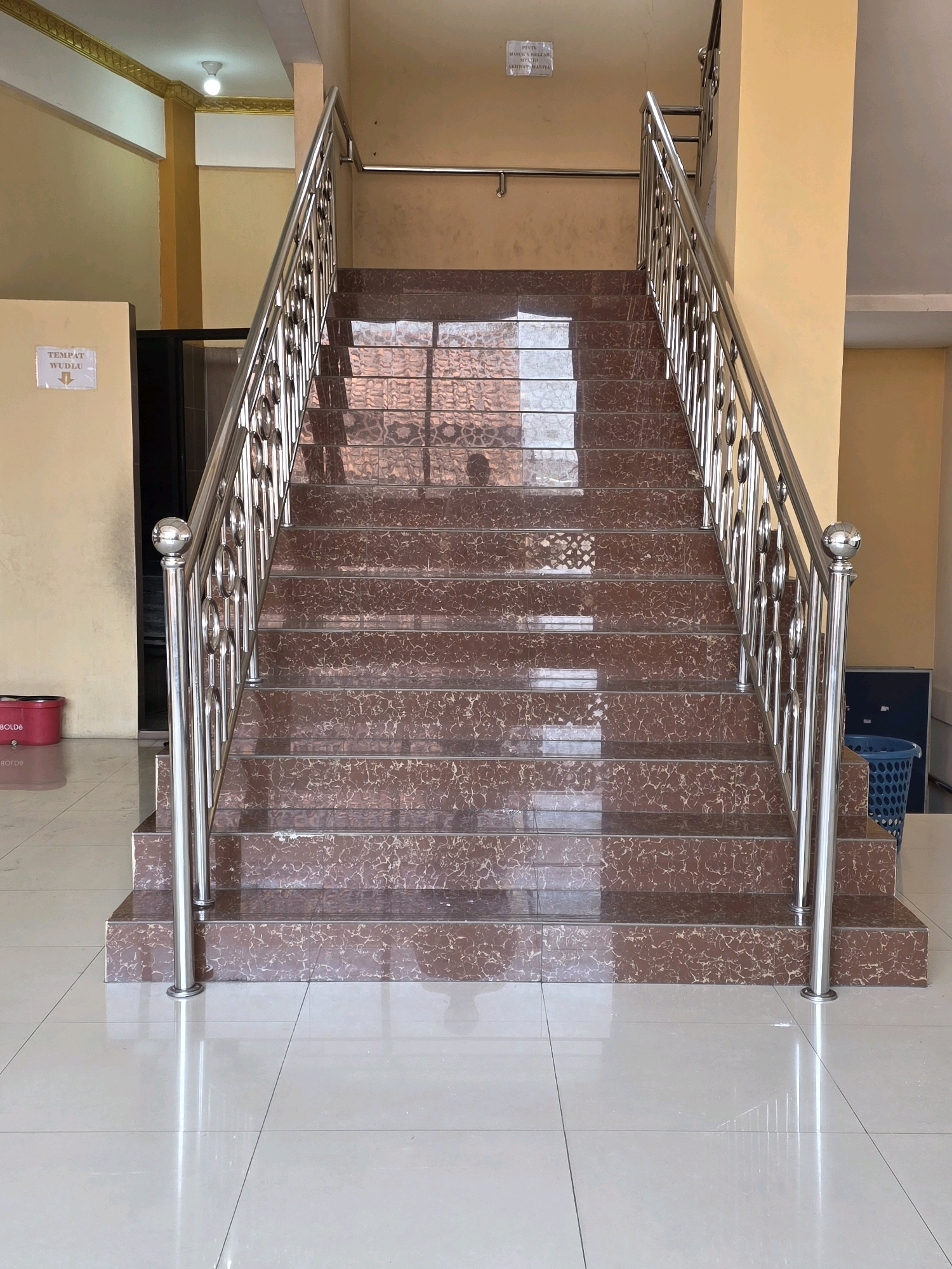 The image shows a modern staircase with polished brown marble steps and a stainless steel railing. The staircase is located indoors, likely in a public building. The walls are painted a light yellow, and the floor is tiled with large, white, reflective tiles. A sign on the left wall reads "TEMPAT WUDLU" with arrows pointing downwards, indicating a place for ablution. A red plastic bucket with the word "BOLDS" is visible on the floor to the left of the stairs. To the right, a blue plastic trash can is partially visible. The lighting suggests an interior space with overhead lights. The overall impression is one of cleanliness and maintenance. This scene is likely from the vicinity of Bojongsoang, Indonesia, given the context provided.