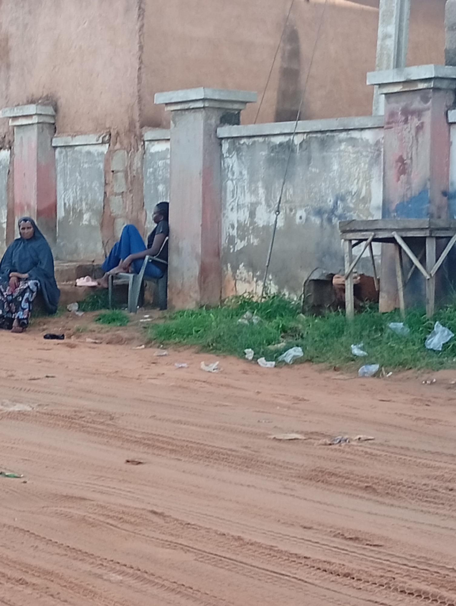 Two individuals are present outside a building with a textured, light brown wall. To the left, a person is seated on a step, wearing a dark headscarf and a patterned skirt. To the right, another person is seated on a dark plastic chair, wearing blue pants and a dark shirt. A light-colored shoe is placed near the steps. The ground in front of the building is covered in reddish-brown dirt with tire tracks. Green grass grows along the base of the wall, and scattered white plastic bags are visible on the grass and dirt. A wooden structure, possibly a table or shelf, is positioned to the right, near some dark, indistinct objects. The overall scene appears to be in an outdoor urban or semi-urban environment in Tumfure, Nigeria.