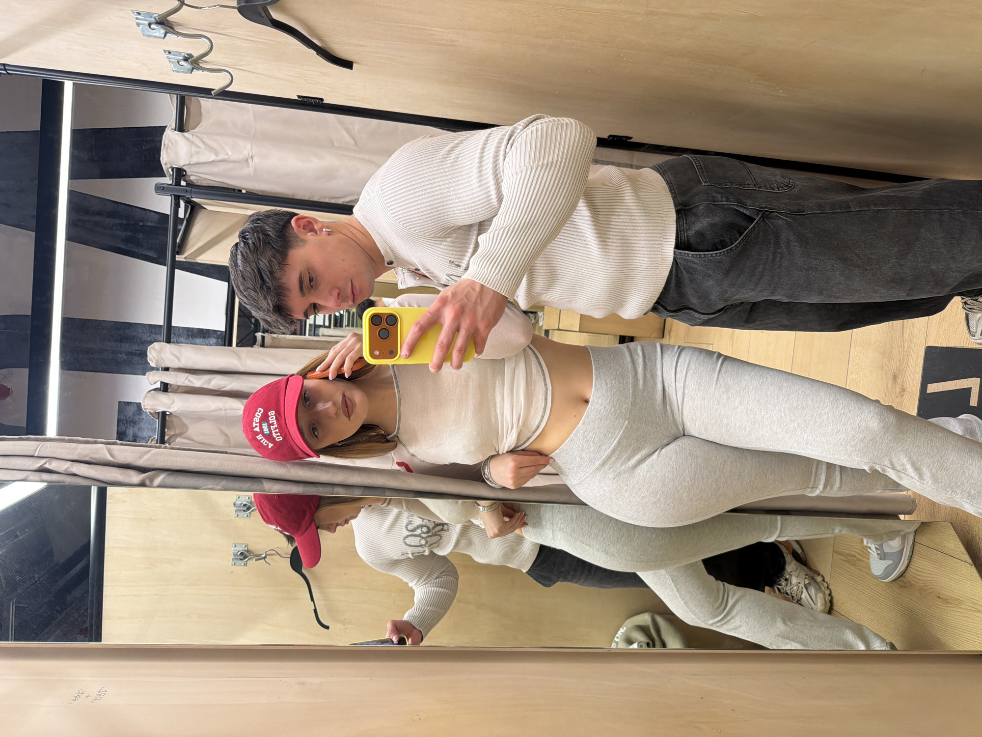 A man and woman are standing in what appears to be a dressing room, taking a picture in the mirror. The man is wearing a cream-colored ribbed sweater and dark jeans. He is holding a yellow smartphone. The woman is wearing a light grey crop top and leggings, with a red baseball cap that says "Buffalo" on it. The dressing room has light wood paneled walls and a mirror with a dark frame. There is a clothing rack with hangers visible in the background. The location appears to be an interior, possibly a retail store dressing room. There is no information to indicate it is in the city of Sofia, Bulgaria.