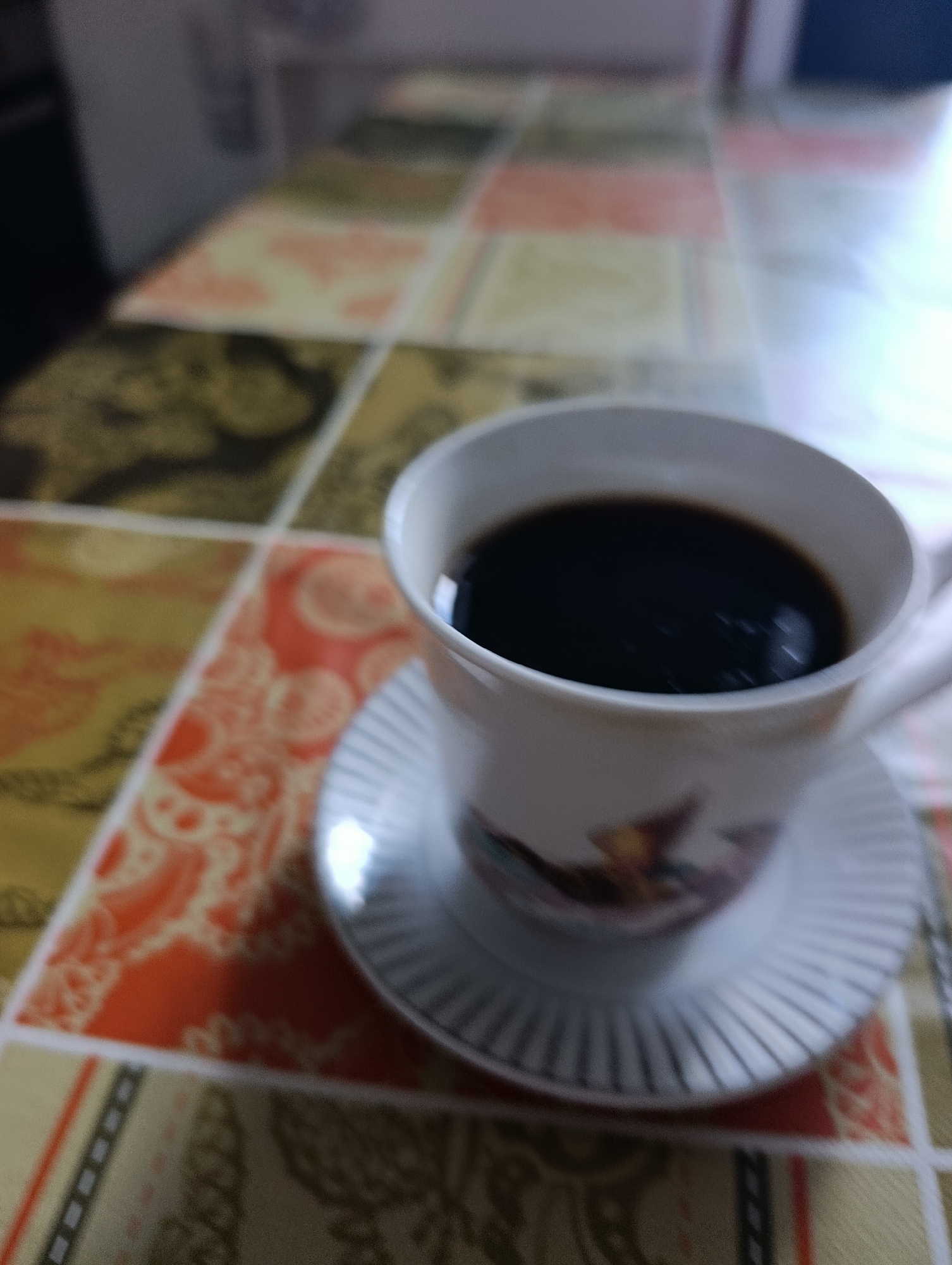 An overhead, slightly angled, close-up shot of a white ceramic coffee cup filled with dark liquid, likely coffee, sitting on a matching white saucer. The cup has a handle, and the saucer has a ribbed pattern. The cup and saucer have a subtle floral or decorative pattern visible on their lower portion. Both are placed on a table covered with a checkered tablecloth featuring squares of various colors and patterns, including orange with white designs, yellow with dark brown designs, and some pink and white squares. The scene appears to be indoors. There is no indication of the city of Setúbal, Portugal, within the image itself.