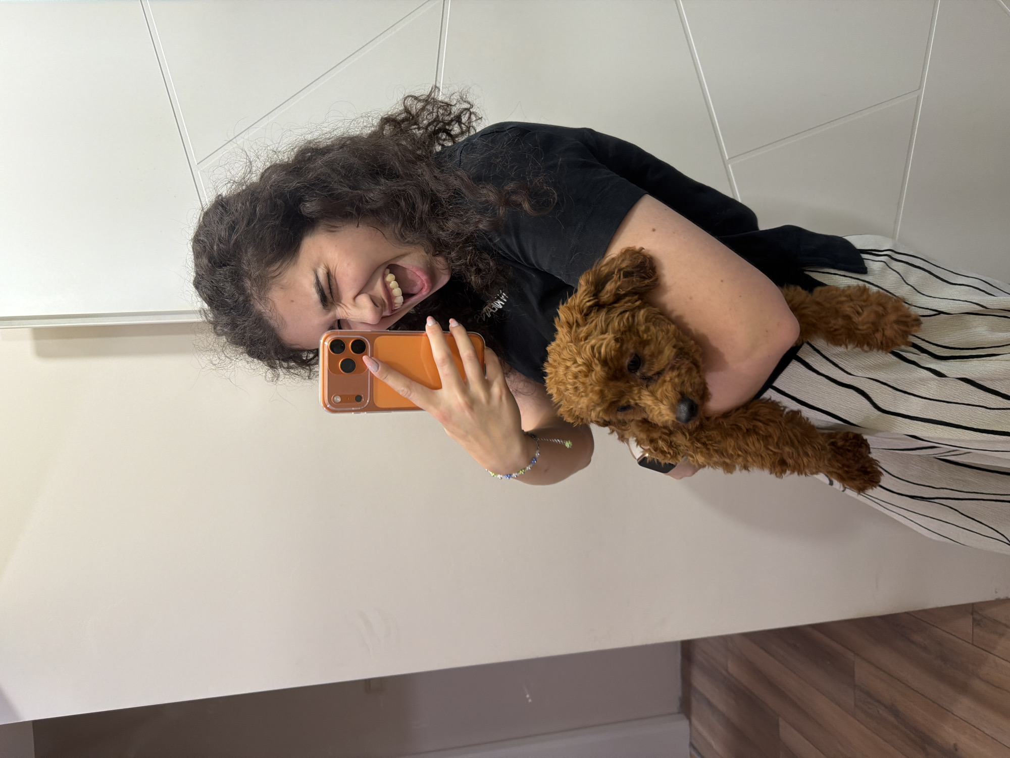 A young woman with curly dark hair is taking a selfie in a mirror, holding a small, fluffy, brown poodle puppy. She is wearing a black t-shirt and white pants with black vertical stripes. The puppy is nestled in her arms, looking down. The background shows a white wall with a geometric pattern and a wooden floor. The scene appears to be indoors, possibly in a residential setting. There is no information in the image to indicate a specific location in Sofia, Bulgaria.