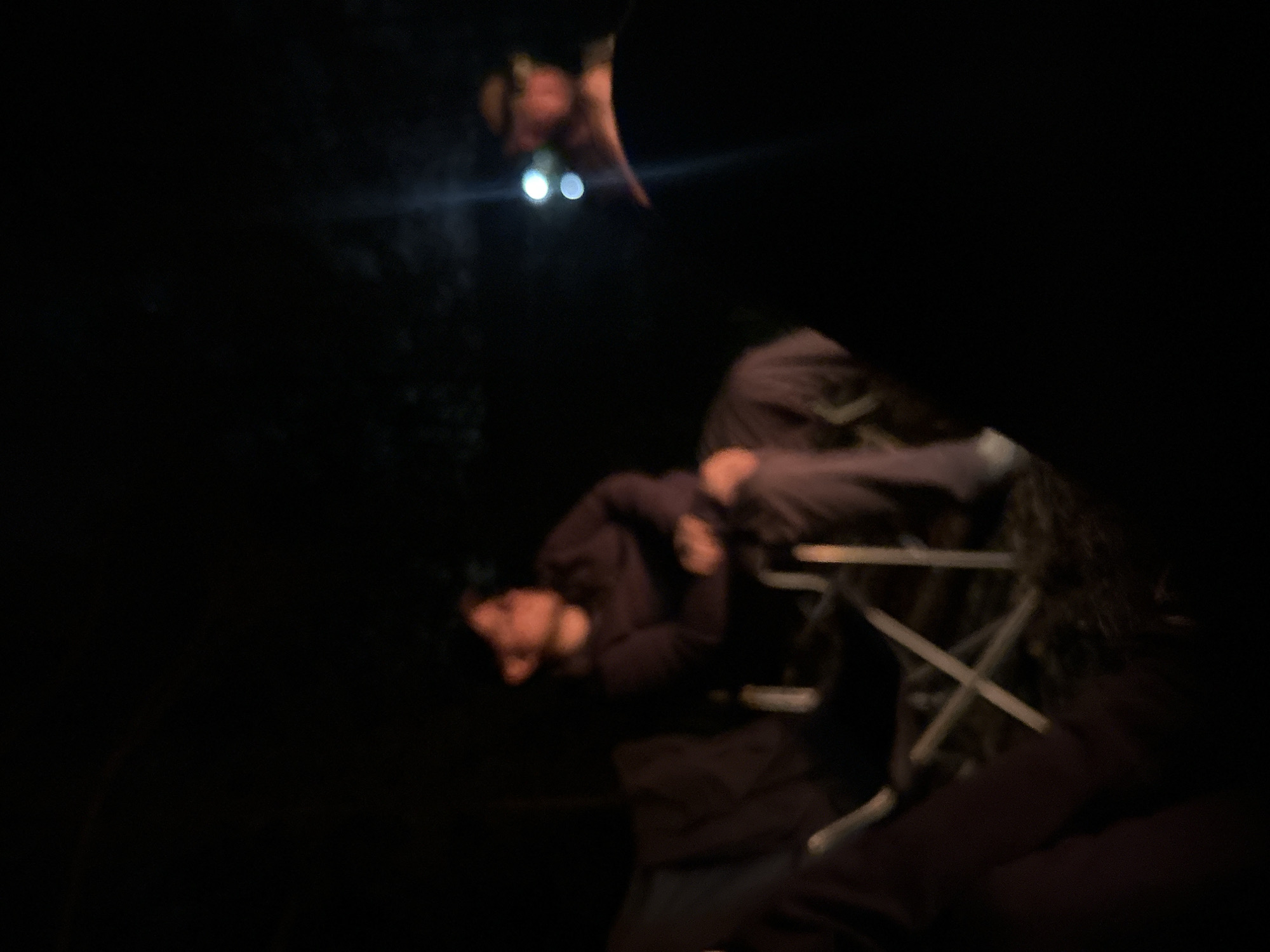 The image, taken in low light conditions, shows two individuals. One person is standing and shining a light downward, while the other is seated in a folding chair. The surroundings are dark. The location is Юзуфоўскі сельскі Савет, Belarus.
