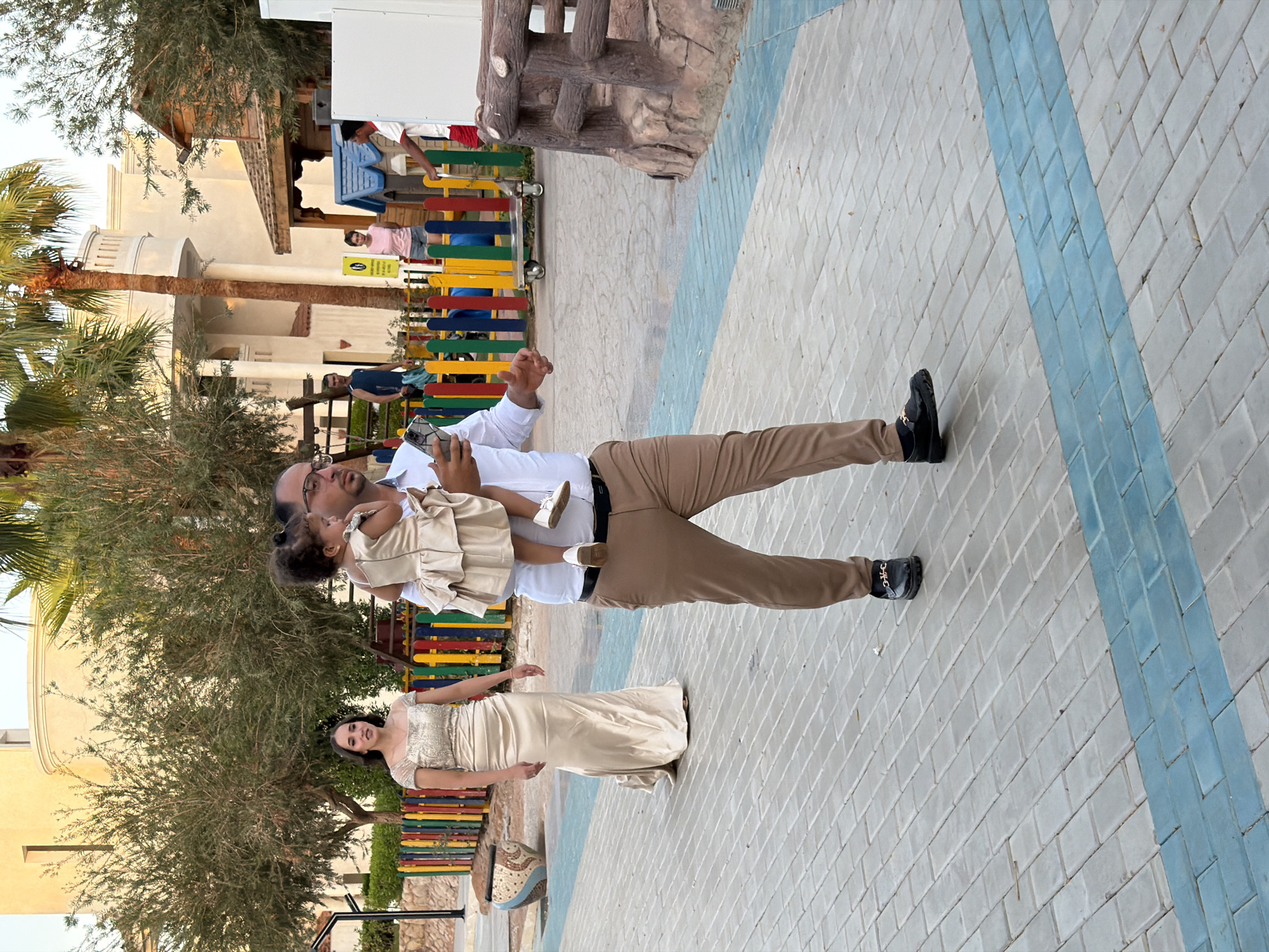 In Sharm El Sheikh, Egypt, a man is carrying a young child while walking on a paved walkway. The man is wearing a white shirt and brown pants, and the child is in a light-colored dress. A woman in a long, gold-colored dress is walking behind them. The background shows buildings, palm trees, and a colorful fence. Other people are visible in the distance, and there is some playground equipment.