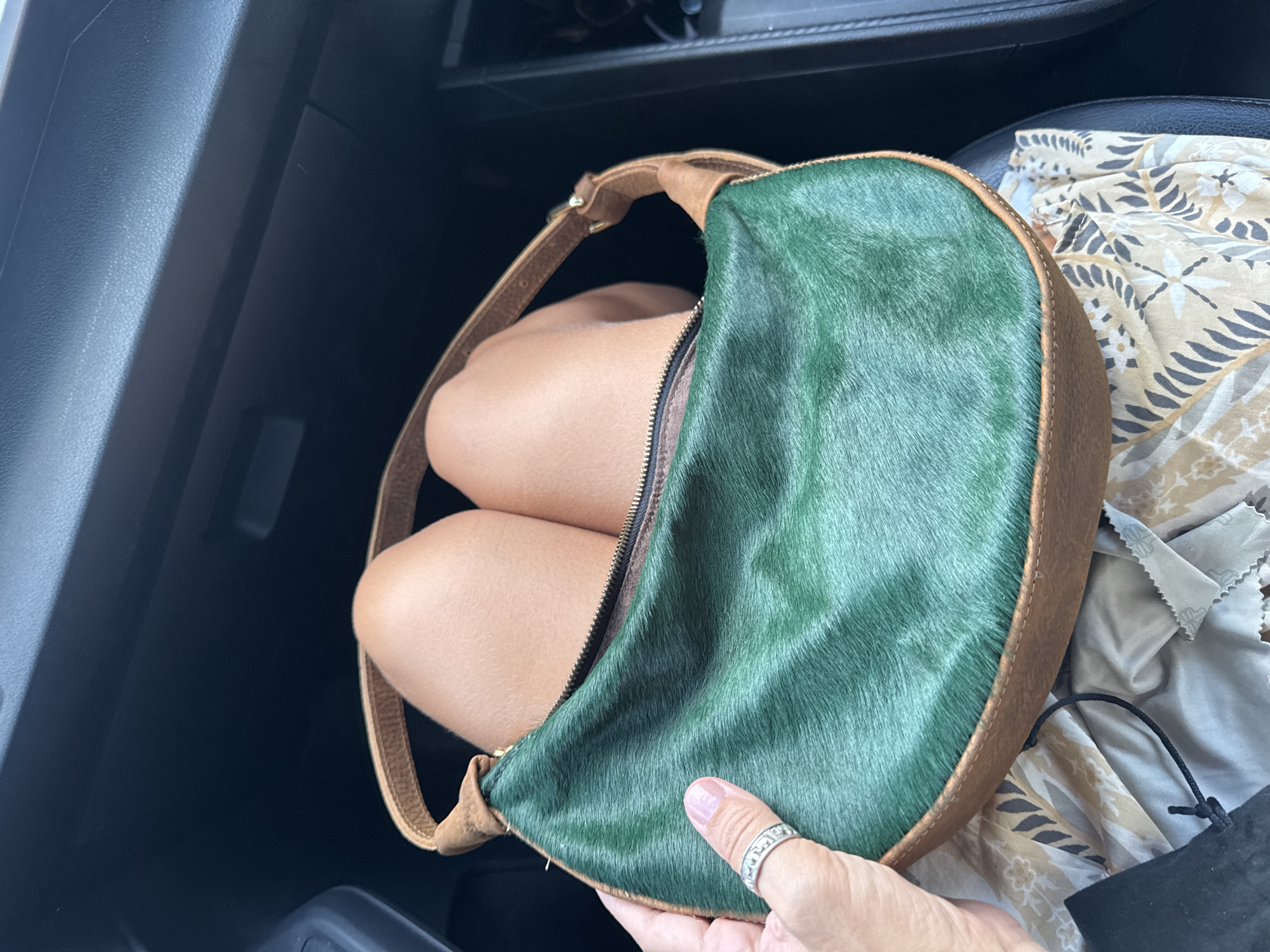 A handbag with a green fur front and brown leather trim is being held in a car. The interior of the car is visible, including parts of the dashboard and seating. Part of a leg is visible inside the handbag. A woman's hand with a ring is holding the handbag. The environment appears to be indoors, inside the car. The location, Kuta Selatan, Indonesia, is specified but not evident from the image.