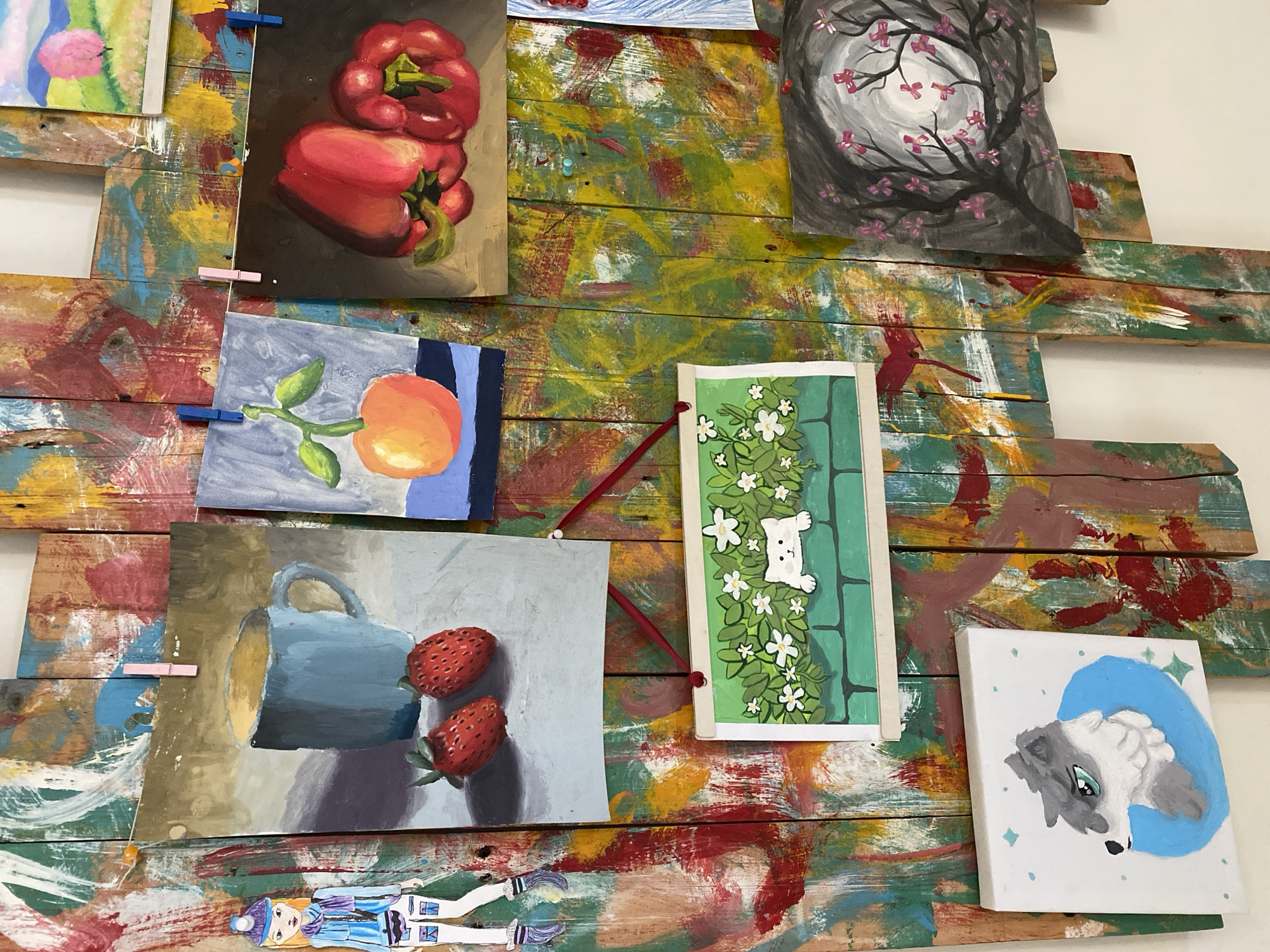 The image depicts a colorful, abstract background of painted wooden planks, likely a wall or a display panel. Affixed to this background are several pieces of artwork, predominantly paintings. On the left, a small, stylized paper doll of a girl in a blue winter outfit is attached.
The paintings include:
* A still life of a blue mug with two strawberries in the foreground.
* A painting of a single orange fruit with green leaves on a branch.
* A close-up of two red bell peppers.
* A rectangular painting of a white cat peeking over a green brick wall, surrounded by white flowers and green foliage.
* A square painting of a wolf's head with gray fur and green eyes, enclosed in a blue circle.
* A painting of a dark, silhouetted tree with pink blossoms against a gray, moonlit sky.
* A small landscape painting with pink trees and a pale blue sky.
The artworks are attached to the wooden planks using clothespins and possibly small nails or pushpins. The overall scene suggests an art display, perhaps in a studio, workshop, or a student's exhibition. There is no visual information in the image to determine its specific location within the city of Đà Nẵng, Vietnam.