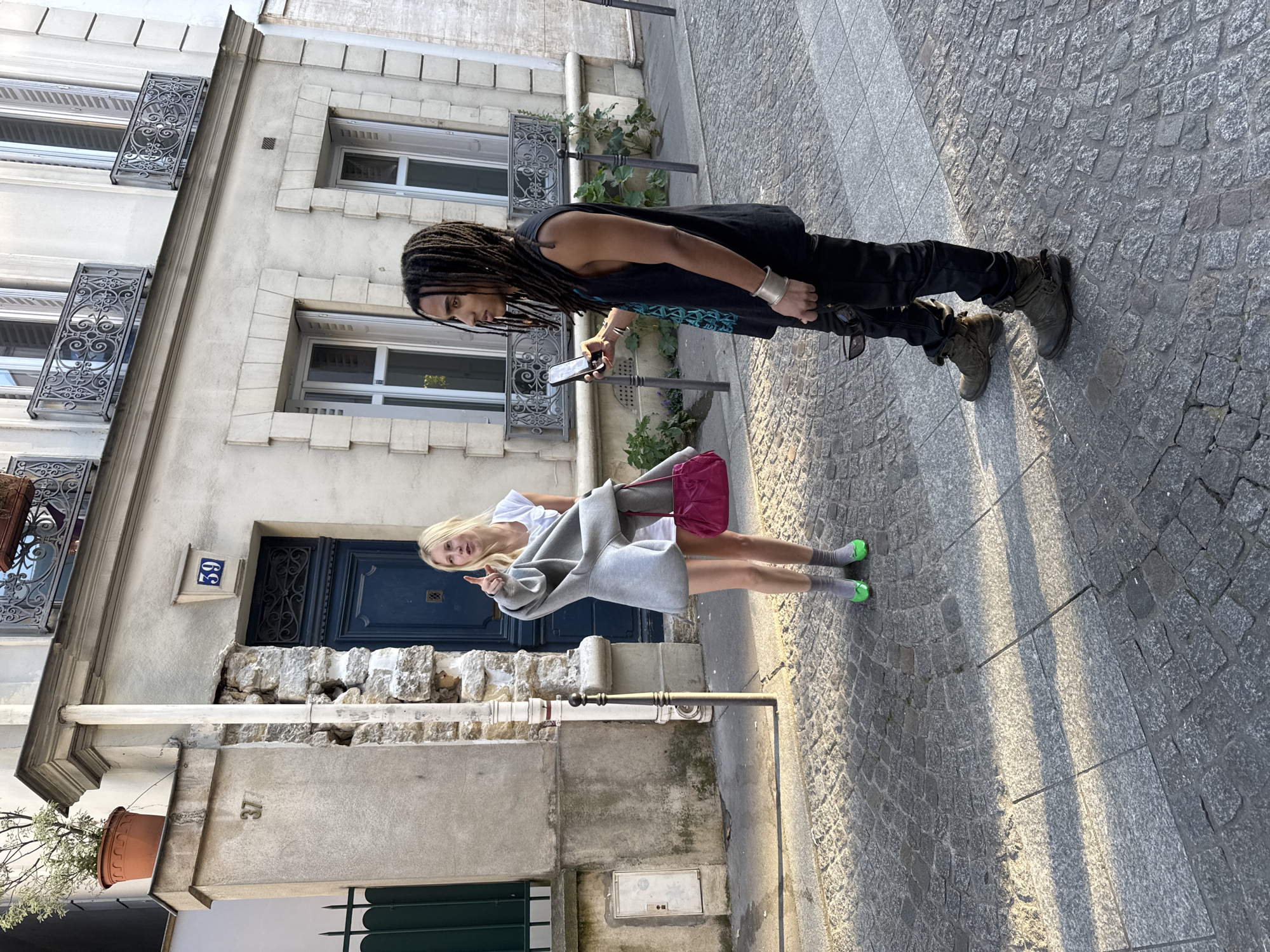 A medium-shot, full-body photograph depicts two individuals standing on a cobblestone street in Paris, France, alongside a building. One individual, positioned at the upper right, is an African American man with dreadlocks, wearing a black tank top, black pants, and dark boots. He holds a smartphone in his hands, seemingly taking a photograph. The other individual is a blonde woman wearing a white top, a gray sweater, and green socks. She gives a thumbs up to the camera. Behind them, the building features white brick, black iron balconies, and dark blue doors. A small, blue and white sign with the number 59 is visible near a doorway. The overall lighting suggests it is daytime.