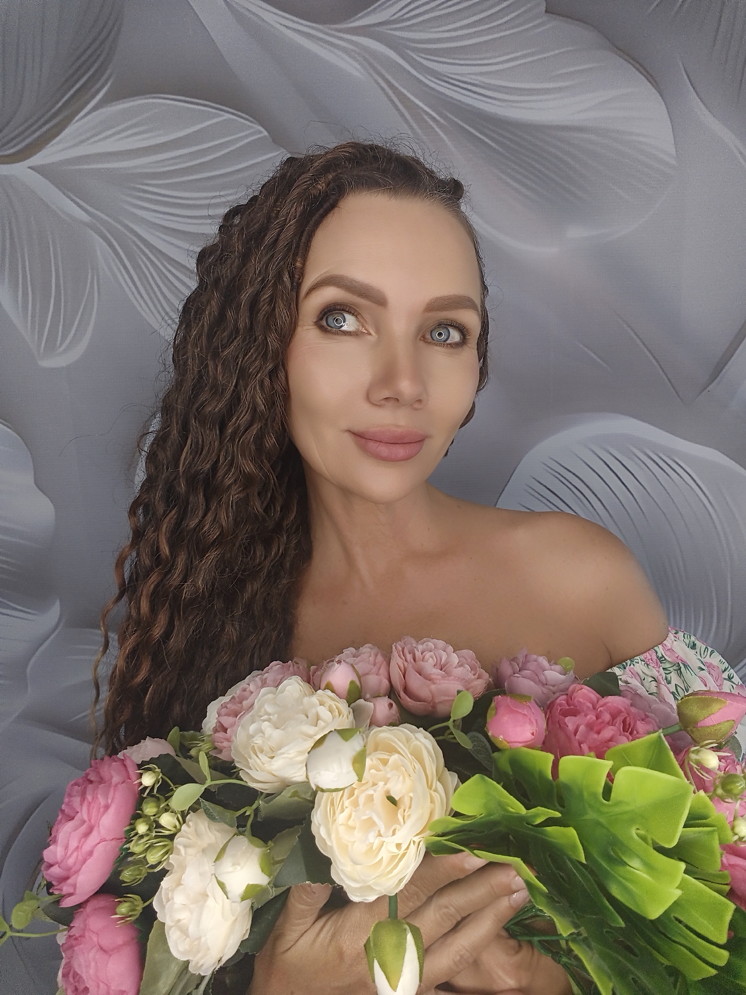 A woman is holding a bouquet of flowers. She has long, curly brown hair and blue eyes. She is wearing an off-the-shoulder dress with a floral pattern. The background is a grey wall with a floral design. The woman is looking to her left and smiling faintly. The flowers are a mix of pink and white roses and green leaves. The lighting appears to be natural, suggesting it is daytime. The overall mood is serene and romantic. There is no visible text in the image.