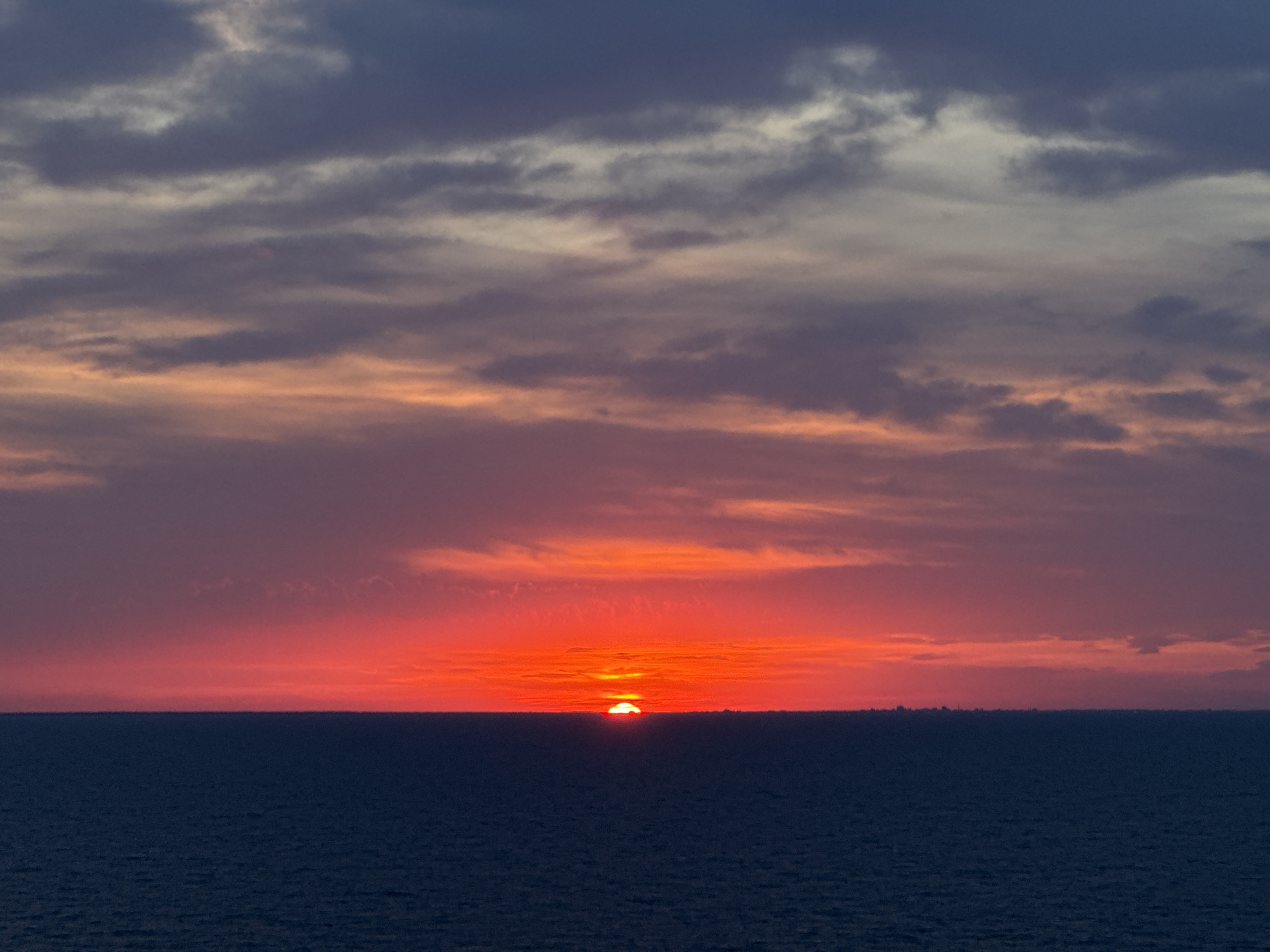 The image depicts a sunset over the sea, with the sun partially visible just above the horizon line. The sky is filled with dramatic clouds painted in vibrant shades of orange, red, and purple, transitioning to darker hues of blue and gray towards the top. The sea below is a deep, dark blue, reflecting the colors of the sky. In the far distance, a faint silhouette of a city skyline can be discerned on the horizon. The scene appears to be a serene and picturesque natural landscape. There are no people, animals, or specific man-made structures clearly identifiable within the frame, aside from the distant city. The overall impression is one of natural beauty and tranquility.