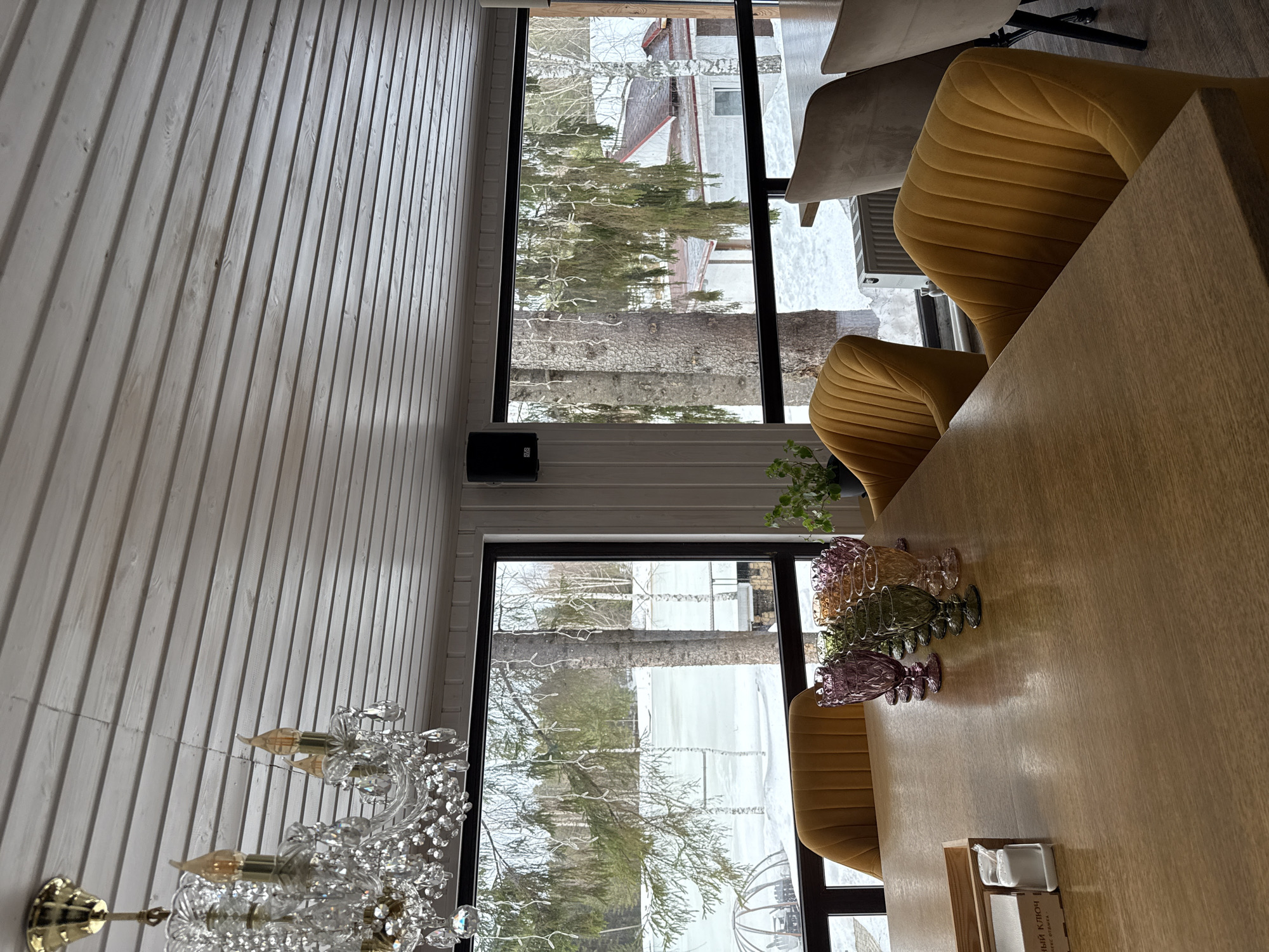 The image depicts an interior scene, potentially a dining area, with a view through a window overlooking a winter landscape, possibly in the vicinity of Votkinsk, Russia. Snow is visible outside, and the leafless trees suggest a winter season.
The interior features a table with several ornate glasses arranged as part of a centerpiece. The table is surrounded by chairs with yellow fabric upholstery. A chandelier is visible, adding to the ambiance. The wall is covered with white planks. The windows have black frames and divide the scene into the interior and exterior.