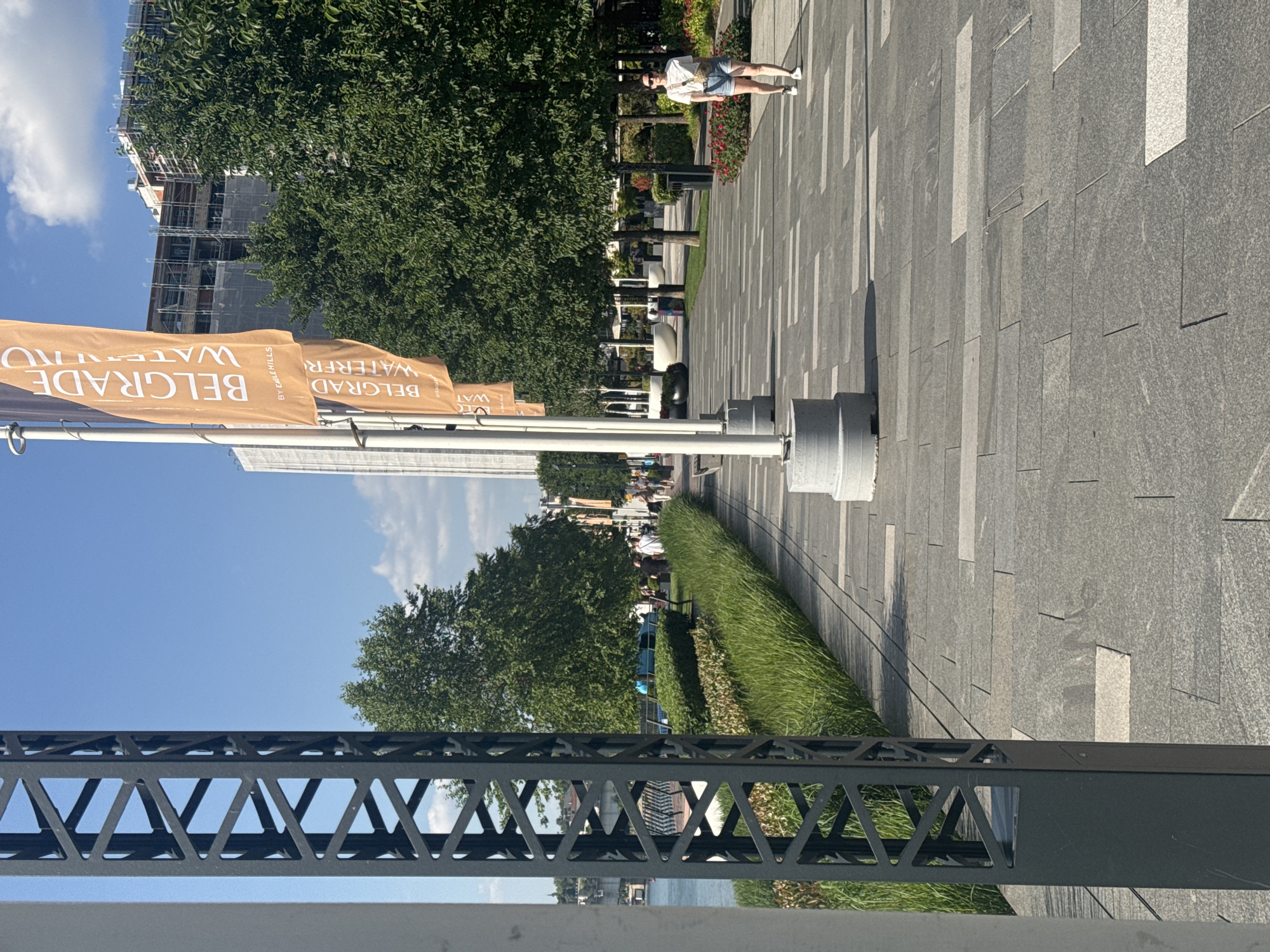 The image captures a sunny day at Belgrade Waterfront in Belgrade, Serbia. In the foreground, a modern, dark metal structural element, possibly a lamp post or part of an art installation, stands to the left, partially obscuring the view. To the right, a wide, paved walkway is laid with large, grey rectangular stones interspersed with lighter grey and white squares. A single woman is walking along this path, dressed in a white t-shirt, denim shorts, and a crossbody bag. She is wearing sunglasses and appears relaxed.
Behind the walkway, a strip of lush green grass and neatly trimmed bushes create a border. Further back, a row of trees with full, green canopies lines the path, providing shade. In the mid-ground, several tall, white flagpoles display orange banners with "BELGRADE WATERFR..." printed in white text. One banner also has "BY EAGLE HILLS" written below it. Behind the trees and flags, a large building is under construction, covered in scaffolding and white netting. A body of water, likely the Sava River, is visible on the far left, with a bridge in the distance. Several other people are seen walking in the background, suggesting a public promenade. The sky is a clear blue with a few scattered white clouds.
