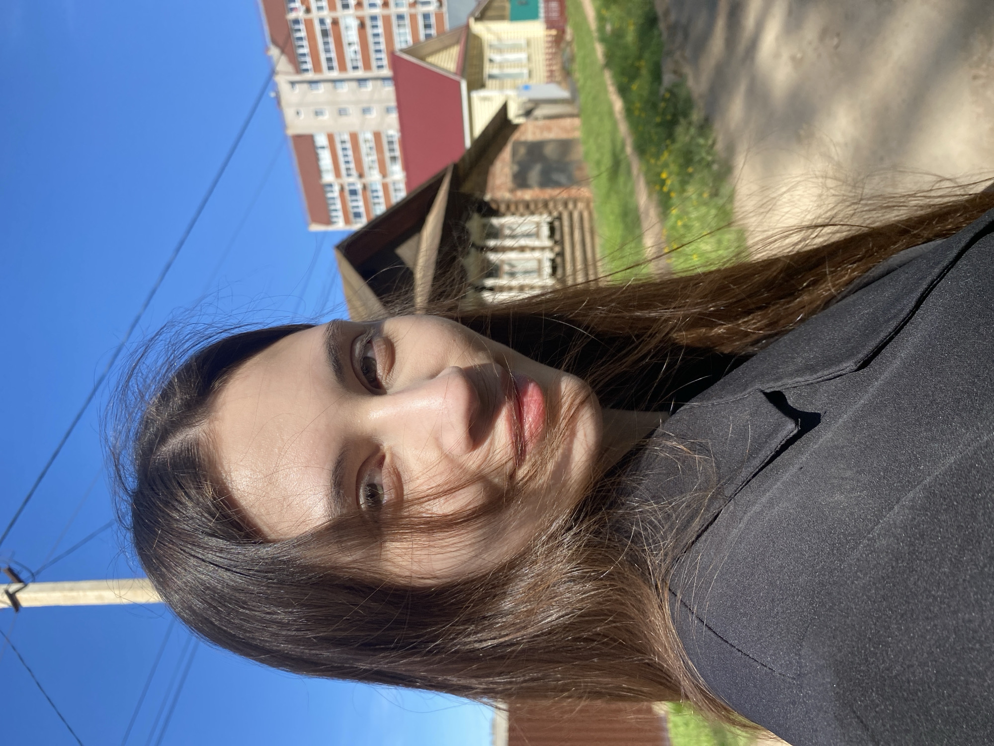 A selfie of a young woman in Votkinsk, Russia. The woman has long brown hair and wears a black top. The photo is taken at an angle, with her face slightly tilted. Behind her are buildings, including a high-rise apartment block with a red roof and a traditional wooden house. A dirt road runs along the background, lined with greenery. The sky is blue.