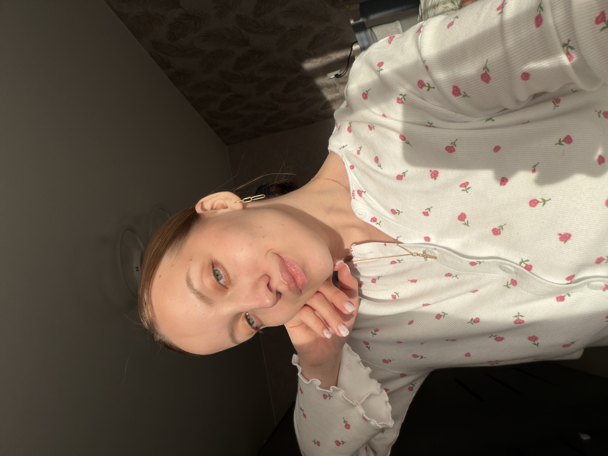 A young woman is pictured in an indoor setting. She is fair-skinned with light brown hair and light-colored eyes. She is wearing a white, long-sleeved top with a pattern of small red roses and a gold necklace. Her left hand is raised to her face. The background includes a dark wall with a light fixture and a patterned ceiling. The location is likely Reshetnikovo, Russia.