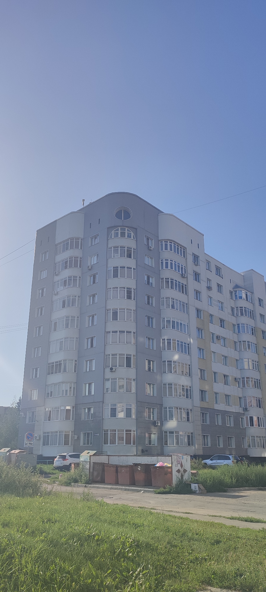 An overcast sky looms over a light gray, multi-story apartment building in Novoaltaysk, Russia. The building has a curved facade with many balconies and windows, some of which are lit. Below the building, a white SUV is parked on the street next to a row of large, reddish-brown trash receptacles. A patch of green grass with some weeds is in the foreground.