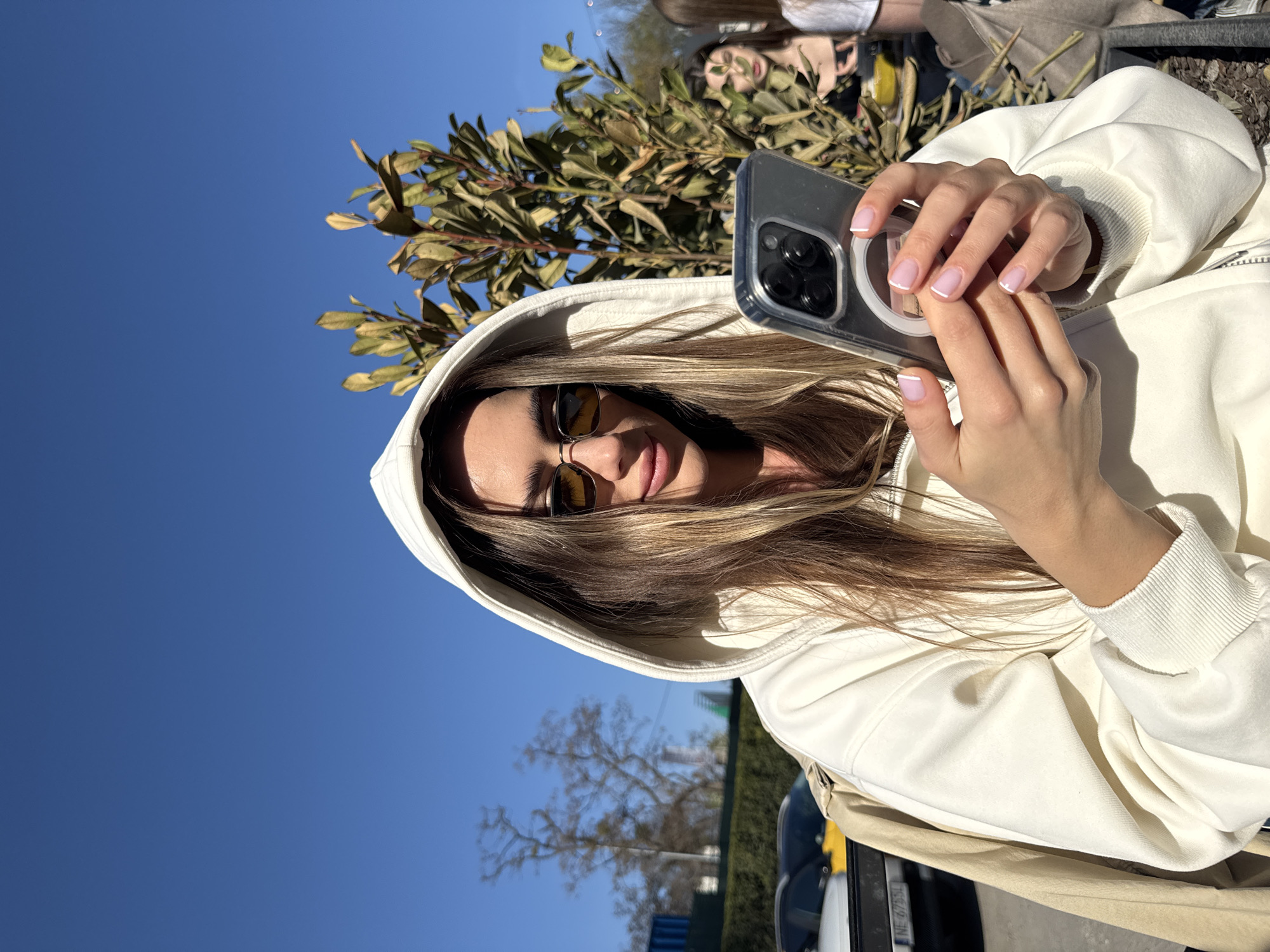 A medium shot of a woman in Warsaw, Poland, holding a smartphone. The woman, wearing a white hoodie, has blonde hair and sunglasses. She is holding the phone in both hands, seemingly taking a selfie or a photo. The background includes green foliage, a clear blue sky, and glimpses of urban elements such as a tree, a car, a blue structure, and other people in the distance.