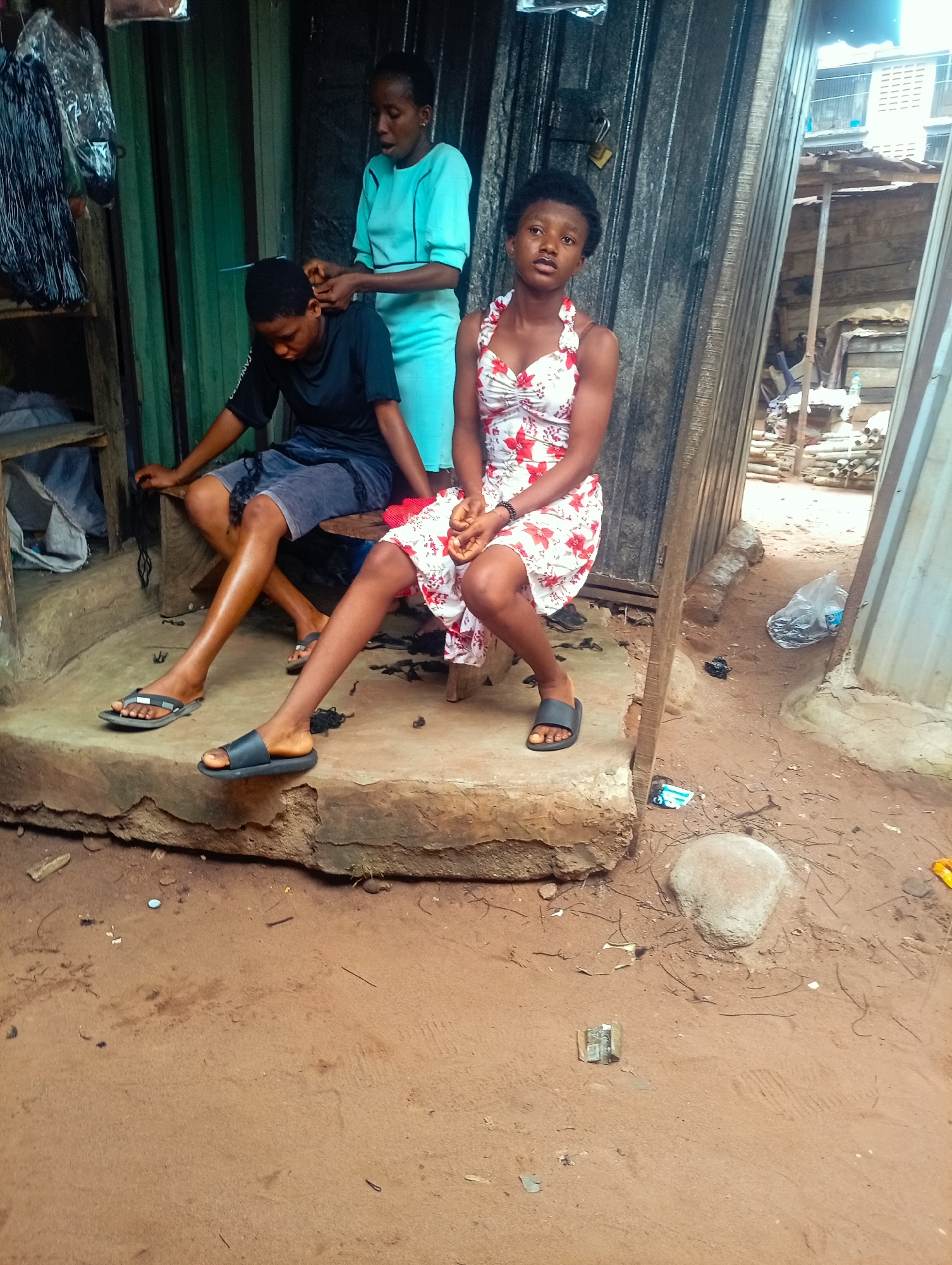 In Nkpor, Nigeria, three young women are present. One woman, wearing a light blue dress, stands and braids the hair of another woman who is seated. The seated woman wears a black t-shirt and blue shorts. A third woman, wearing a floral print dress, sits to the right of the other two. The setting appears to be an outdoor area with a corrugated metal structure in the background, possibly a shop or stall. The ground is sandy and there are some scattered debris.