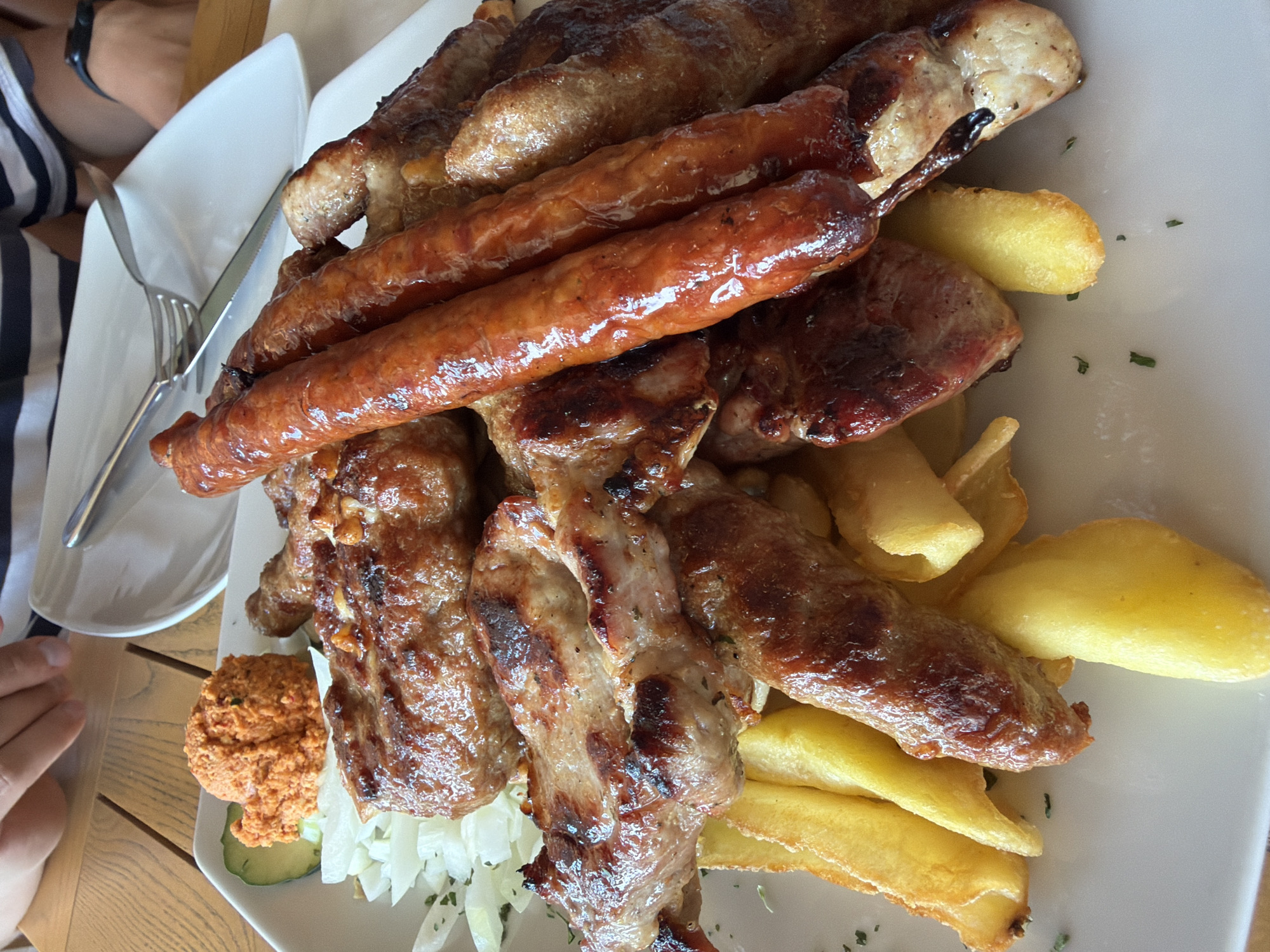 An overhead shot shows a plate of grilled meats and french fries, likely at a restaurant in Bigor, Montenegro. The plate, filled with a generous portion of food, is set on a wooden table. The meal includes several types of grilled meat, including sausages and what appear to be small meat patties or kebabs. French fries are piled around the meats, and a small mound of reddish paste, possibly ajvar or a similar relish, is visible on the side of the plate next to a slice of cucumber and some shredded lettuce. A person's hands and arm, wearing a striped shirt and a watch, are visible in the upper left corner, reaching towards the food. Another person's legs are visible in the upper right corner. A fork and knife are placed on a white plate in the background.