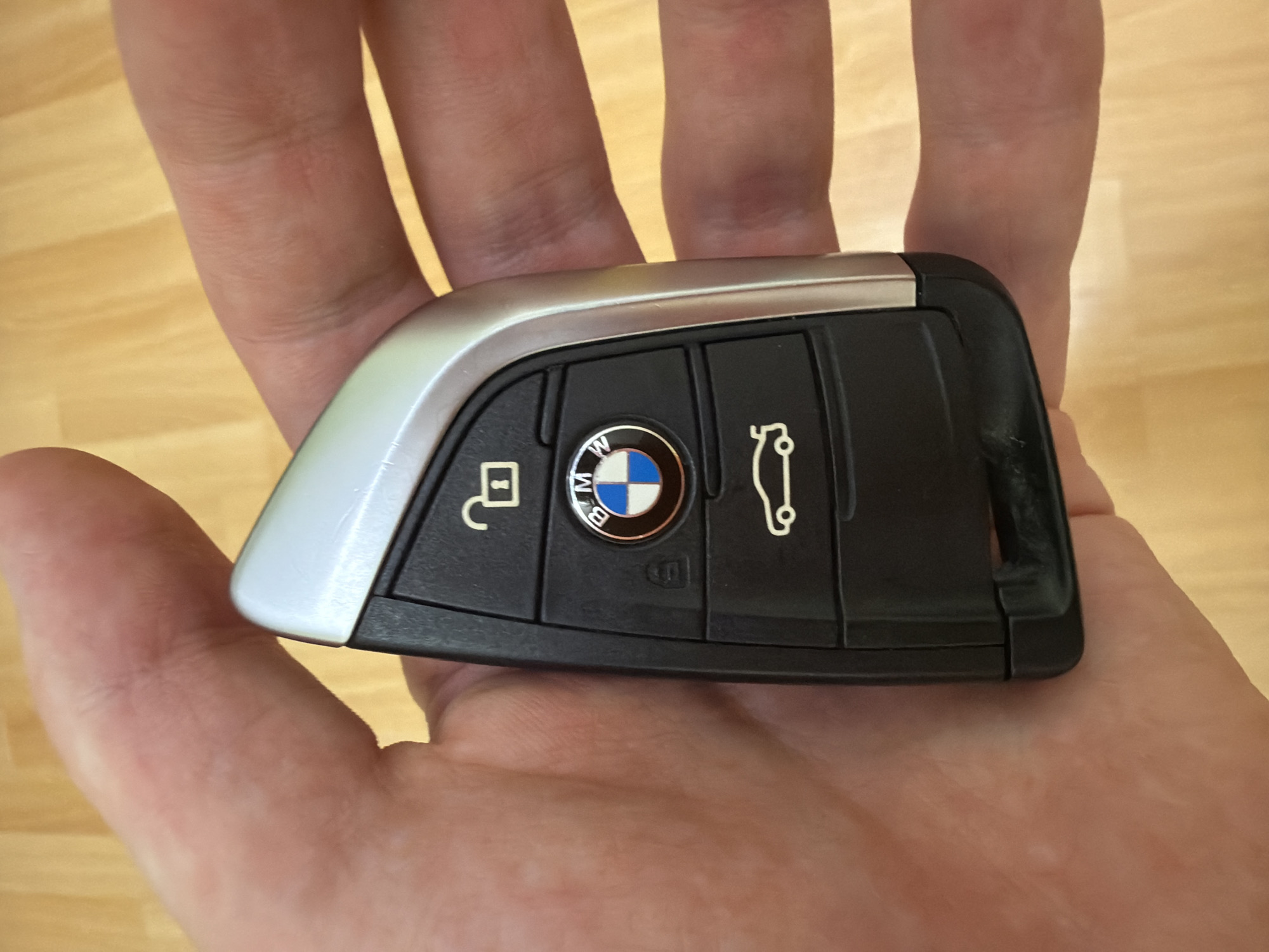 A person's hand holds a black and silver BMW car key. The key has three buttons with icons: an unlocked padlock, the BMW logo, and a car silhouette. The hand and key are positioned over a wooden surface, likely a table or floor. The lighting suggests an indoor environment.