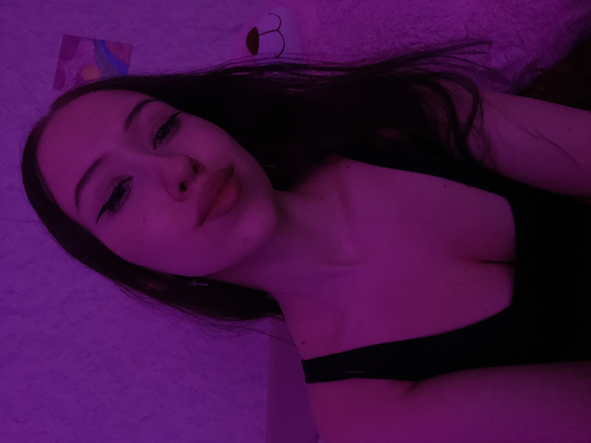 This is a close-up, medium shot photograph taken indoors, likely in a bedroom. The scene is dominated by a soft, purple ambient light, possibly from a neon or LED light source. The background consists of a textured wall, on which a small, abstract painting featuring a rainbow and sunset is hung. To the right, a portion of a fluffy white teddy bear is visible.
The main subject is a young woman with long, dark hair, wearing a black tank top. Her face is angled slightly towards the camera, and she has winged eyeliner. She is wearing a small cross earring. The lighting casts a vibrant purple hue over the entire image, illuminating her skin and features. The focus is on her upper body and face, with the background providing context for an intimate, personal space. The photograph appears to be a selfie, given the angle and framing. There is no indication of the city of Votkinsk, Russia, within the image itself.