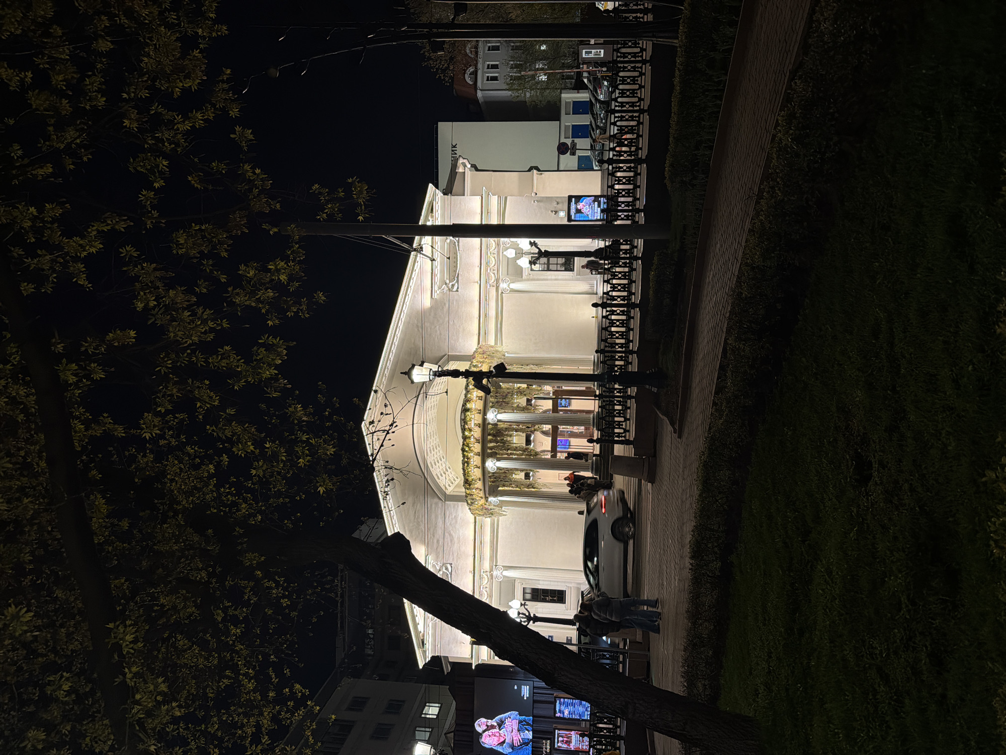 This media file captures a nighttime urban scene in Moscow, Russia, with the image rotated 90 degrees clockwise from its natural orientation. The central focus is a grand, brightly illuminated building, possibly a theater or public institution, featuring classical architecture with columns, an ornate facade, and a curved portico adorned with greenery or garlands. Its pale structure glows warmly against the dark night sky.
In the upper portion of the image (originally the left), dark tree branches with clusters of light green leaves are visible against the black sky, with faint distant lights. Below the central building (originally the right), a paved pathway runs alongside a dark, dense hedge or bushes, where a few individuals are seen.
On the left side of the image (originally the top), distant buildings with some lit windows are visible, and on one, the Cyrillic word "БАНК" (BANK) is clearly readable. On the right side of the image (originally the bottom), the thick trunk of a large tree dominates the foreground. Near its base, multiple brightly lit display screens are visible, one prominently showing an image of an older man and woman in a warm embrace.
In front of the main building, a white car, possibly a taxi, is present, and several people are standing on the sidewalk, suggesting a public gathering or activity. Street lamps provide additional illumination. The time of day is night, with artificial lights creating a vibrant and well-lit atmosphere.