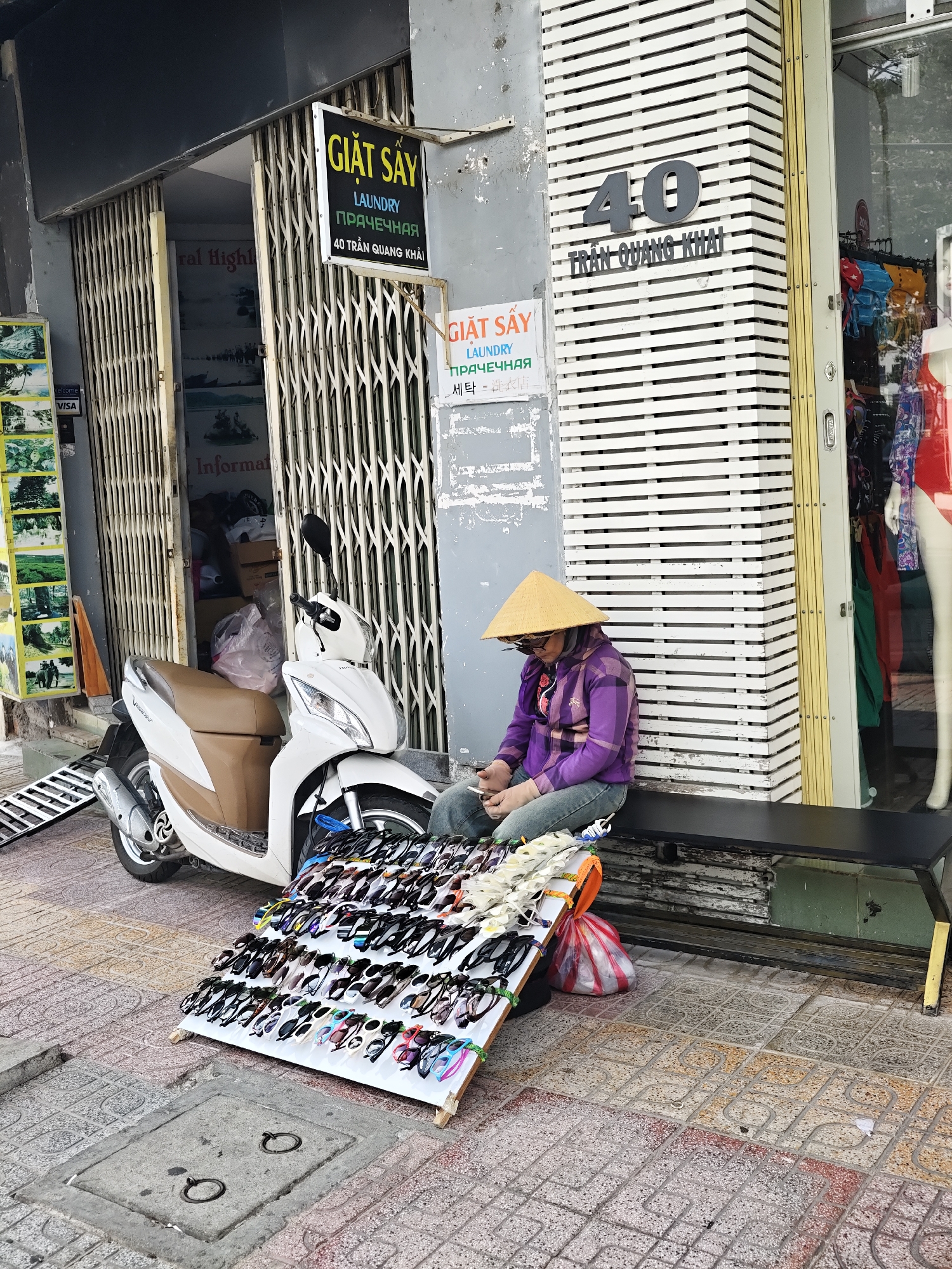 A person wearing a conical hat and sunglasses sits on a black bench in front of a store in what appears to be Nha Trang, Vietnam. The person is dressed in a purple jacket and blue jeans and is looking down at a mobile phone. In front of them, a display rack is filled with sunglasses for sale. A white scooter is parked to their left. The store has a striped facade and a glass window displaying clothing. Signs on the building indicate "GIẶT SẤY LAUNDRY" in Vietnamese, Russian, and Korean. The ground is paved with textured tiles. The scene suggests a street vendor selling sunglasses outside a laundry business. The time of day appears to be daytime, with bright sunlight casting shadows.