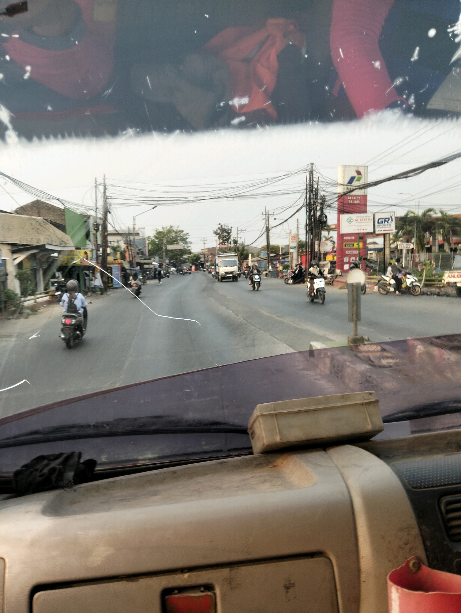 The image captures a street scene in Semarang, Indonesia, viewed from inside a vehicle, likely through the windshield. The foreground reveals the dashboard of the vehicle, with a light-colored box and a red bucket-like object. The windshield itself has several white, irregular lines, possibly from damage or reflections.
The middle ground shows a moderately busy street with several motorbikes and a white truck. On the left side of the road, there are buildings, some appearing to be shops or houses with awnings. A person in a blue shirt and helmet is riding a black motorbike. Further down the road, other motorbikes are visible, some with passengers.
The background features more buildings, utility poles with numerous wires crisscrossing, and trees. A prominent sign for "PERTAMINA" (an Indonesian state-owned oil and gas company) with "AL FATEEH" and "GR SPORTS" also visible on other signs. The sky is overcast, suggesting it might be late afternoon or an earlier hour. The overall impression is a typical urban street scene in Indonesia.