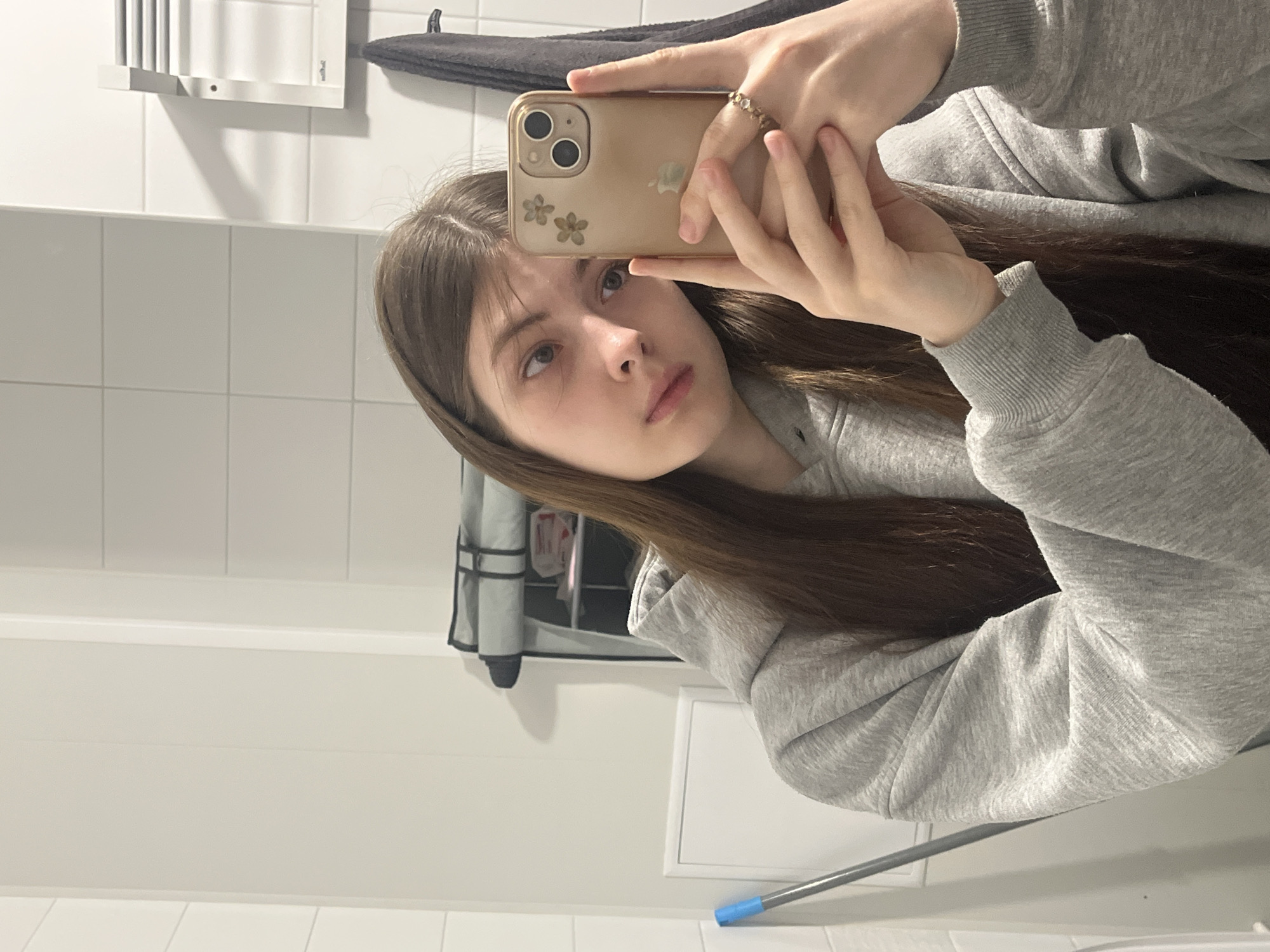 A young woman with long brown hair is taking a selfie in a bathroom. She is wearing a gray hoodie. The bathroom has white tiled walls and features a towel rack and other bathroom fixtures. The location is not discernible from the image and cannot be determined to be in Saint Petersburg, Russia.