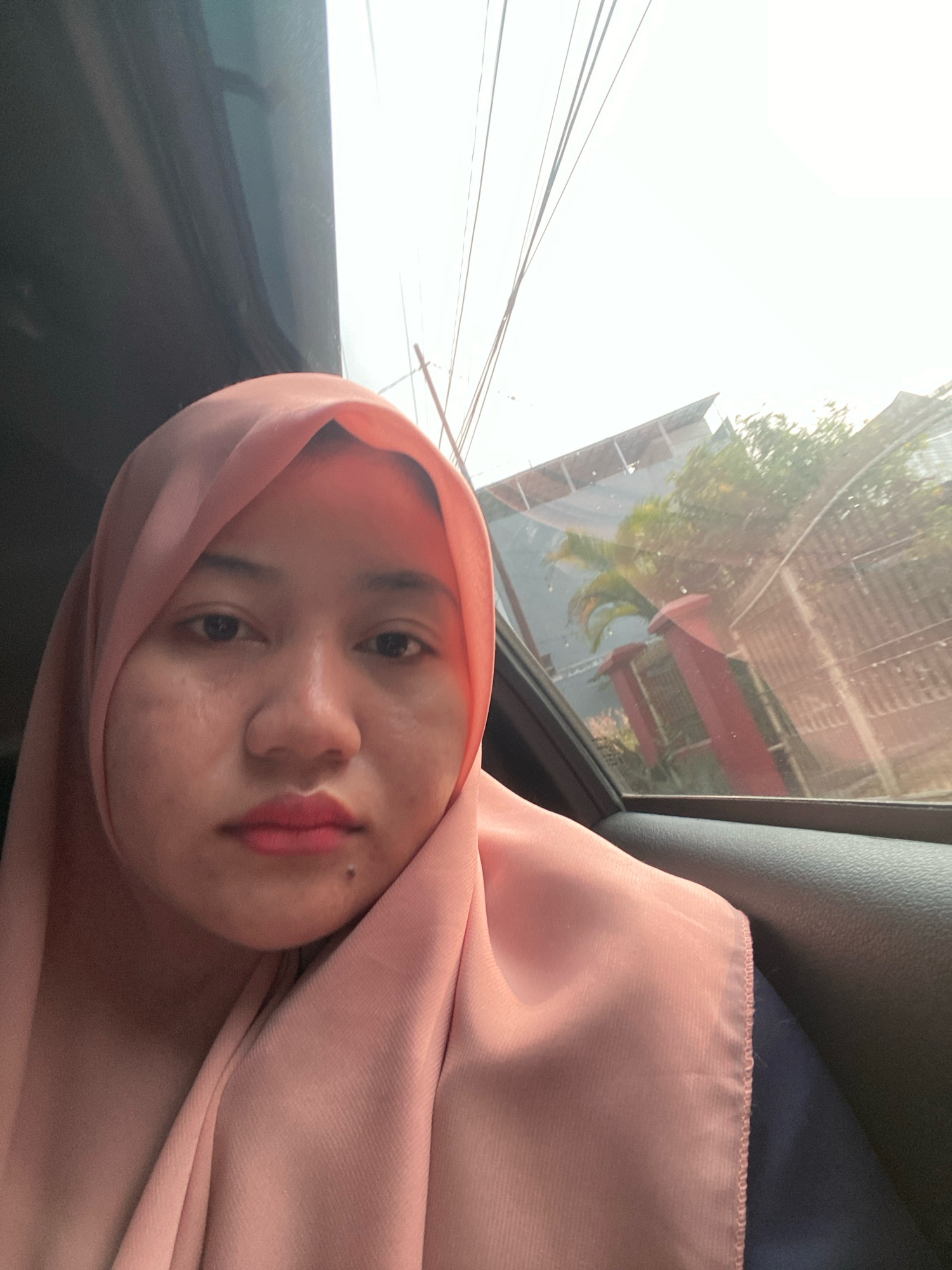This is a selfie taken from inside a vehicle, with the subject, a young woman wearing a pink hijab and pink lipstick, positioned in the foreground. The woman is looking directly at the camera. Through the car window on the right side of the image, a blurry outdoor scene is visible, featuring a fence, some vegetation, and a building with a slanted roof. The sky is bright and overcast. The interior of the car is visible on the left side of the image, showing a dark surface, likely the car's interior paneling. The image appears to have been taken during daylight. The woman's location within the city of Banjarbaru, Indonesia, is not discernible from the visual information provided in the image itself.