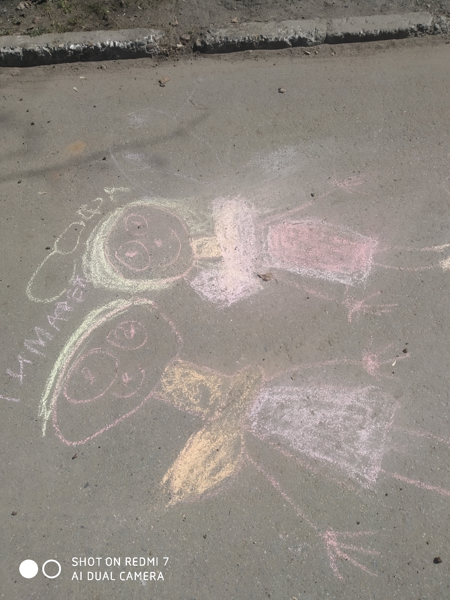 The image shows two chalk drawings of cartoon-like figures on asphalt. The drawings are located on the street in the city of Chaikovsky, Russia. The drawings consist of two circular heads with eyes, bodies, and limbs. The drawings are colorful, using chalk in shades of pink, yellow, green, and white. There is handwriting in Russian next to one drawing. The surrounding asphalt is textured and shows signs of wear. A sidewalk is visible at the top of the image.